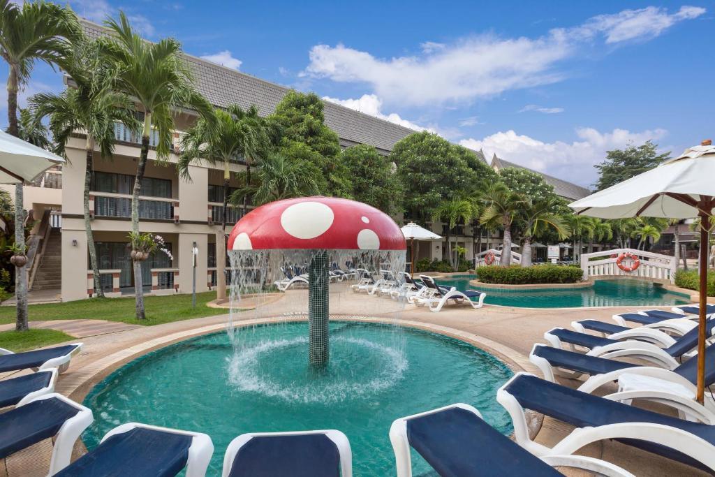 Red and white mushroom-shaped water feature spraying in shallow turquoise baby pool surrounded by lounge chairs at tropical resort.