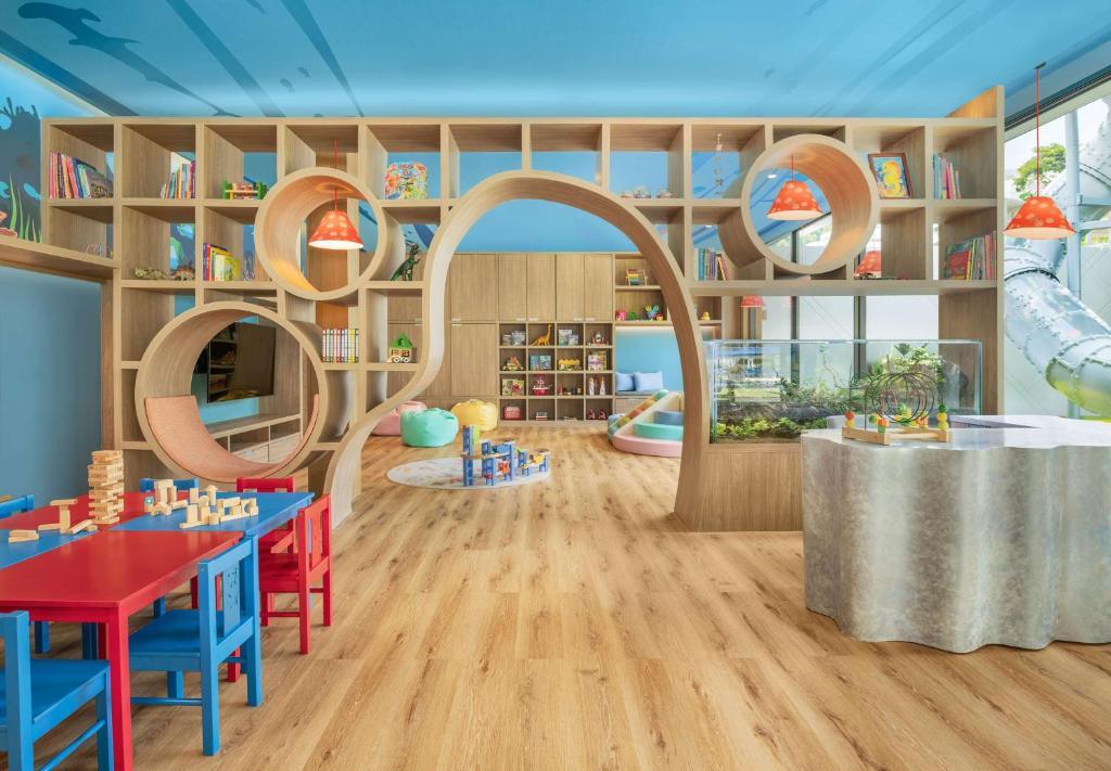 Bright kids club interior with wooden circular play tunnels, red and blue activity table, shelving with books and toys, and orange pendant lights.