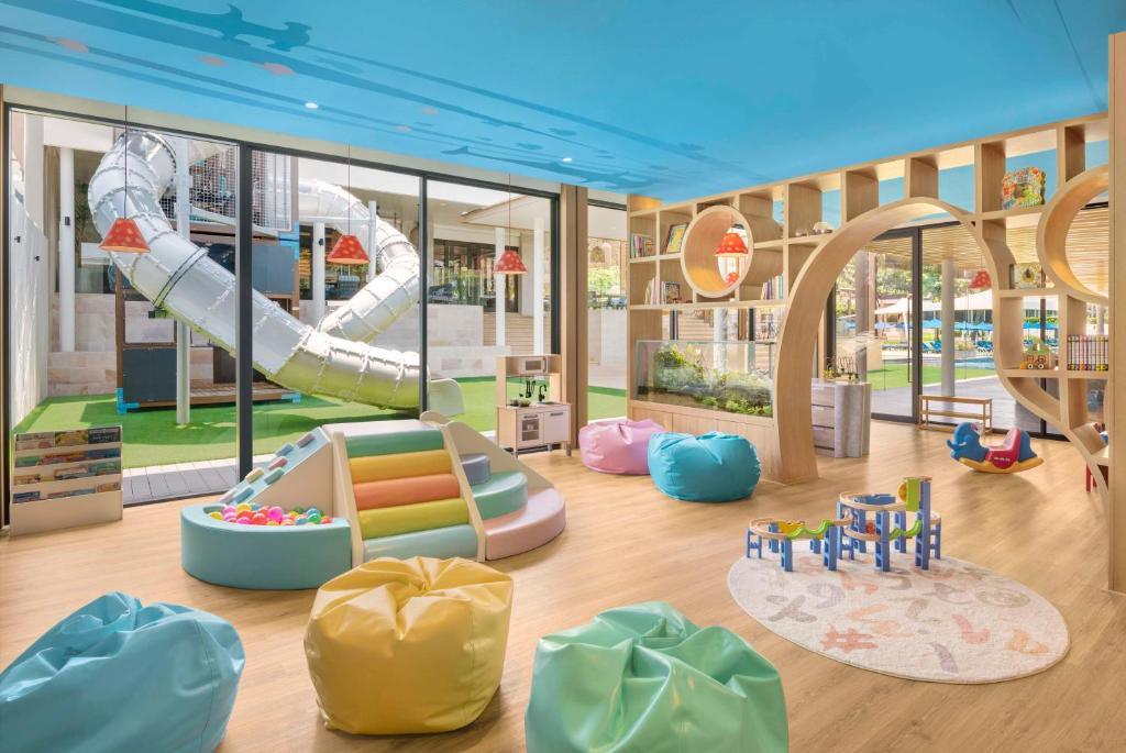 Bright kids club interior with colorful bean bags, wooden climbing structure, tube slide, and toy blocks on wood flooring.