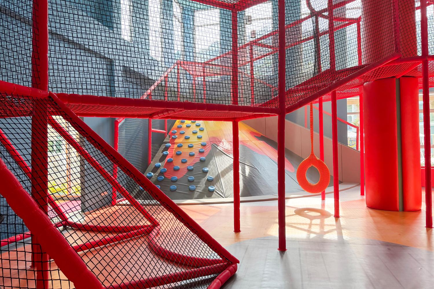Bright red indoor kids club with climbing wall, slide, and net play structures filled with natural light.