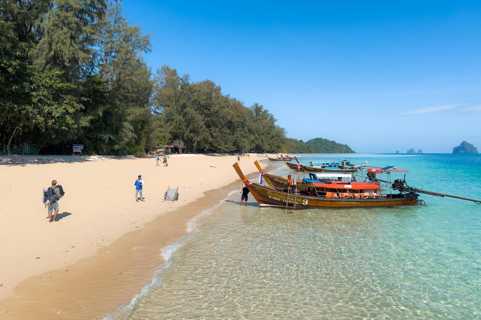 Tropical beach at Koh Kradan, Thailand, with turquoise water, wooden longtail boats, white sand and visitors swimming and walking.