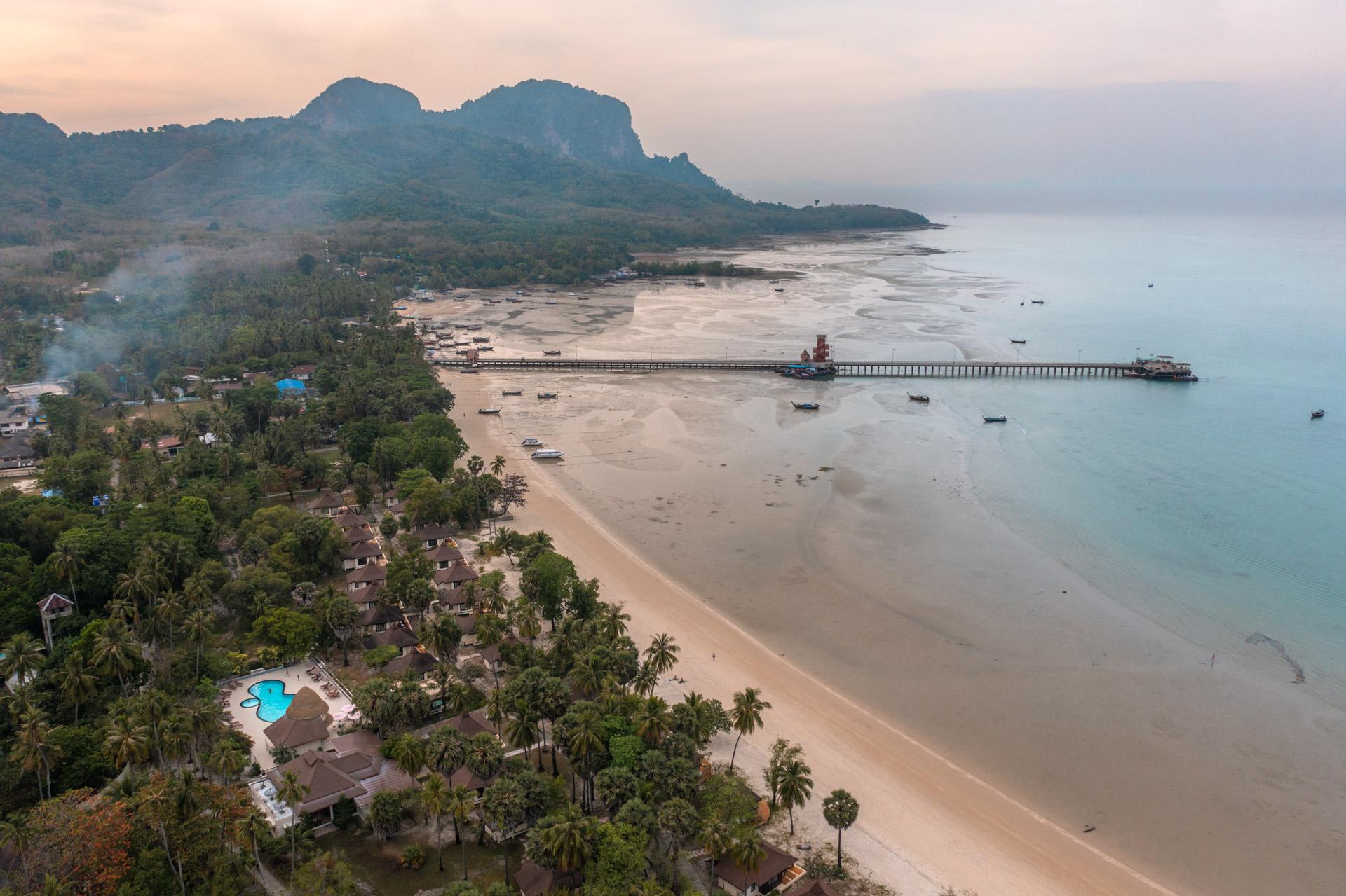 Aerial view of Koh Mook's sandy beach, resort with blue pool, limestone cliffs, pier and turquoise waters dotted with boats at dusk.