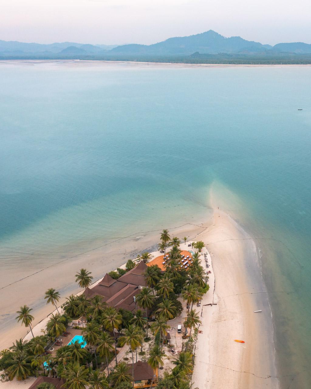 Aerial view of Koh Mook's sand spit with palm trees, resort buildings, turquoise water and mountains in the background.