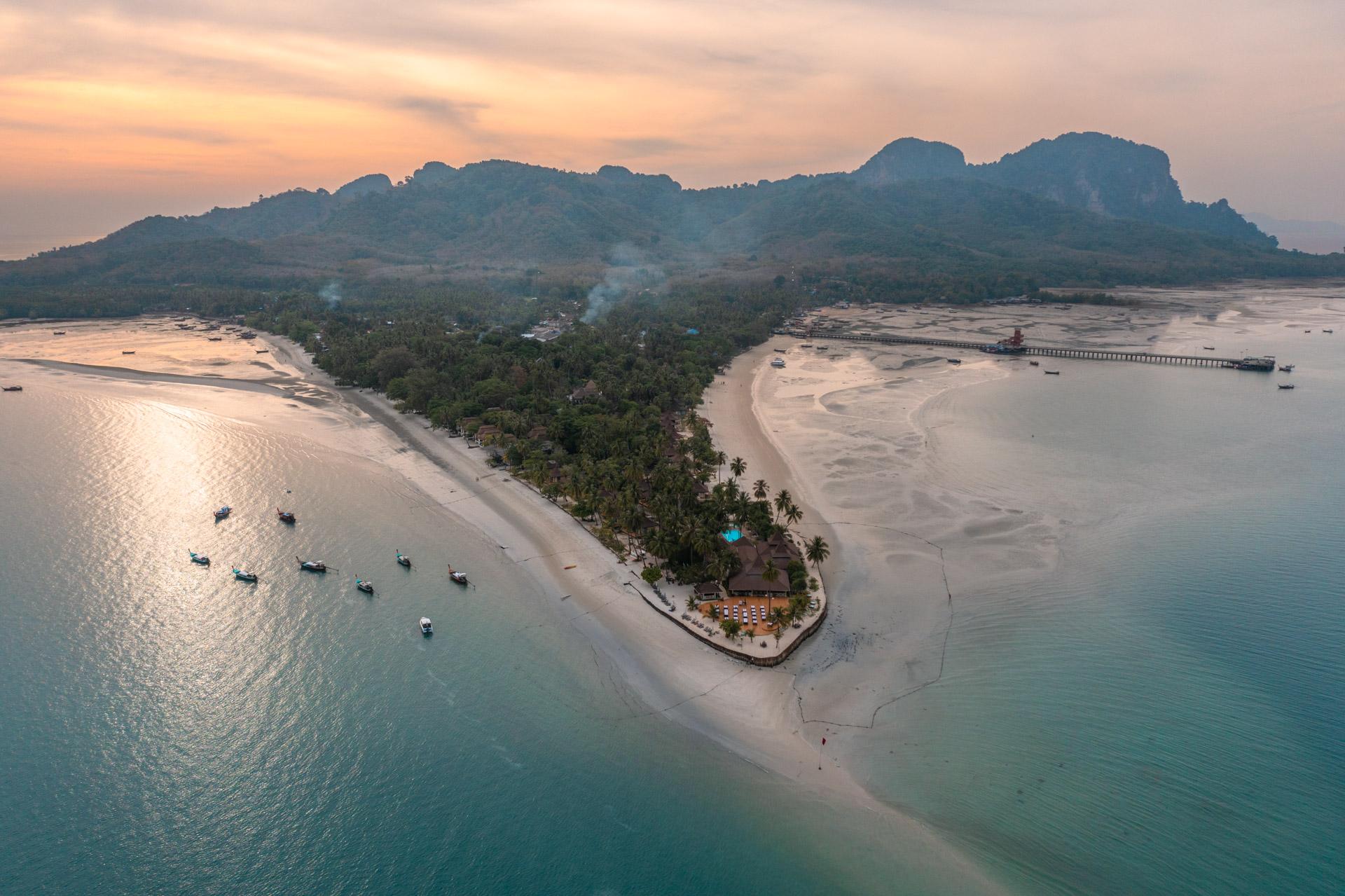Aerial sunset view of Koh Mook island with limestone cliffs, sandy beaches, resort and longtail boats in turquoise waters.