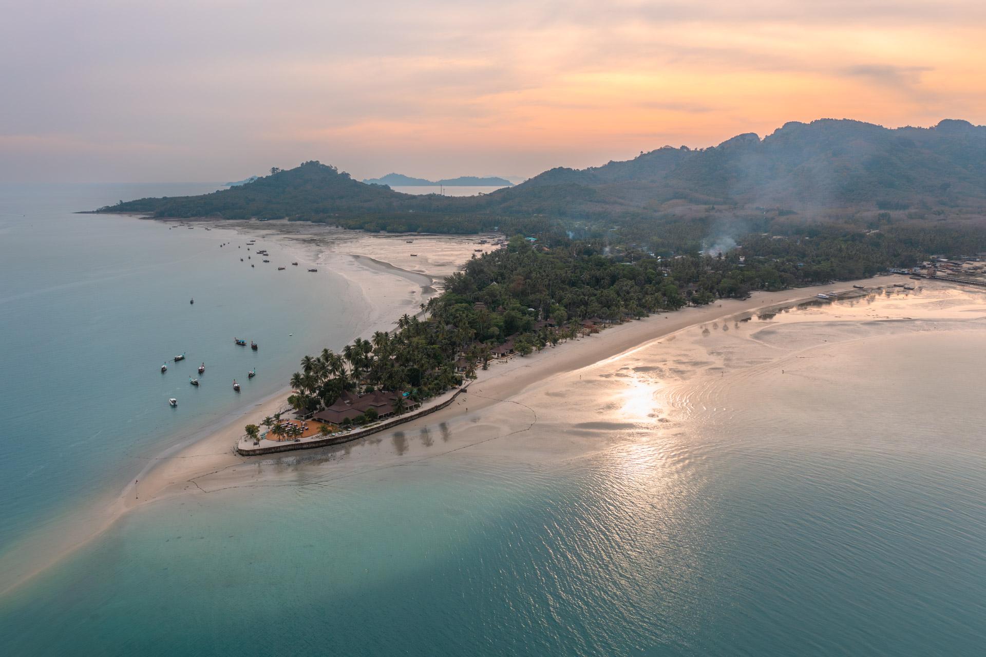 Aerial view of Koh Mook island at sunset with turquoise waters, sandy beaches, palm trees, moored boats, and forested mountains in background.