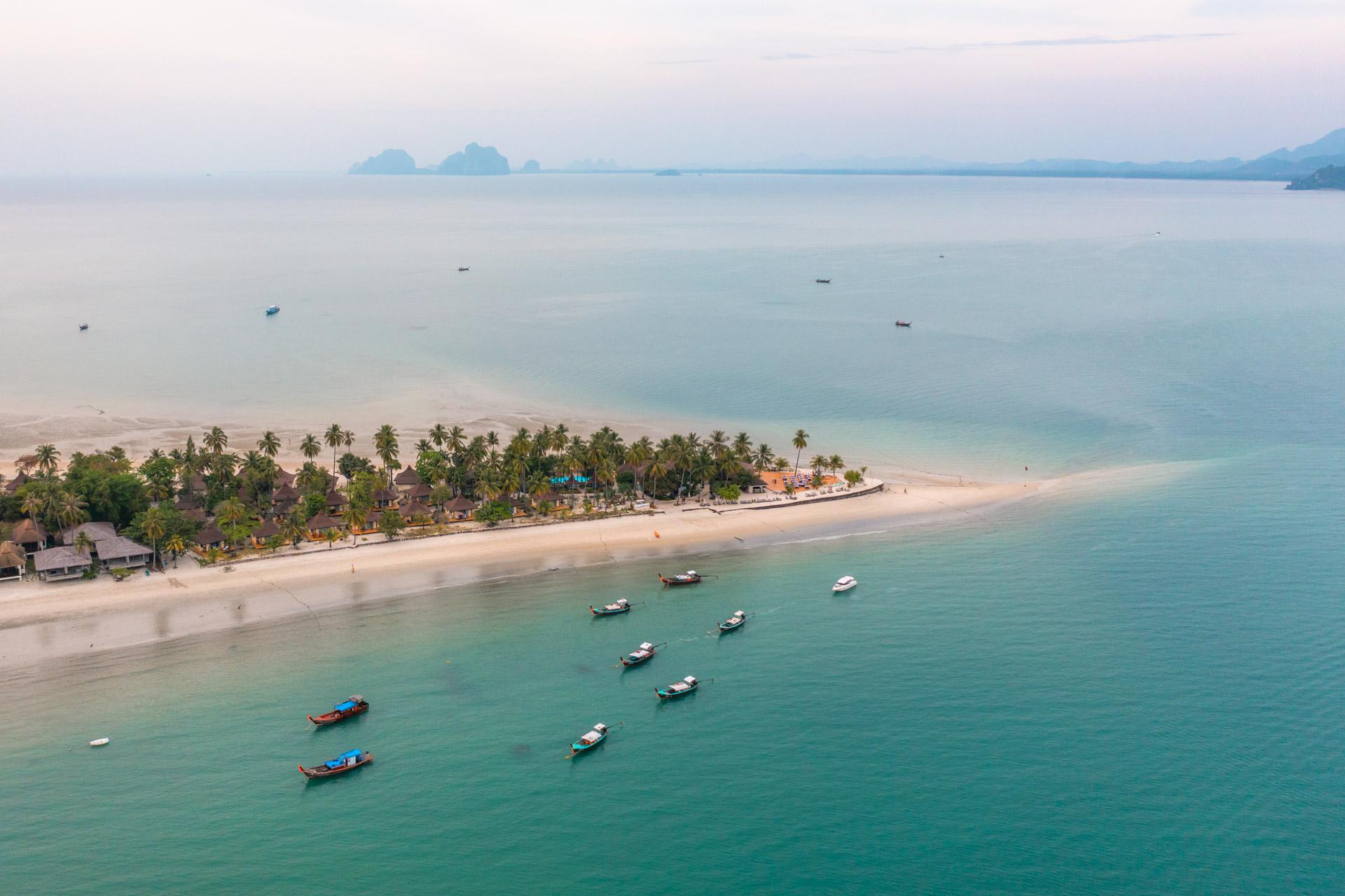 Aerial view of Koh Mook beach with turquoise waters, traditional fishing boats, palm trees and limestone islands in distance.