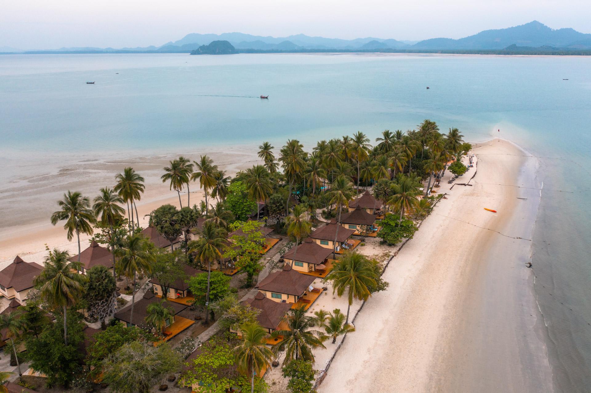 Aerial view of Koh Mook island resort with sandy beach, palm trees, wooden bungalows, turquoise water and distant limestone mountains.