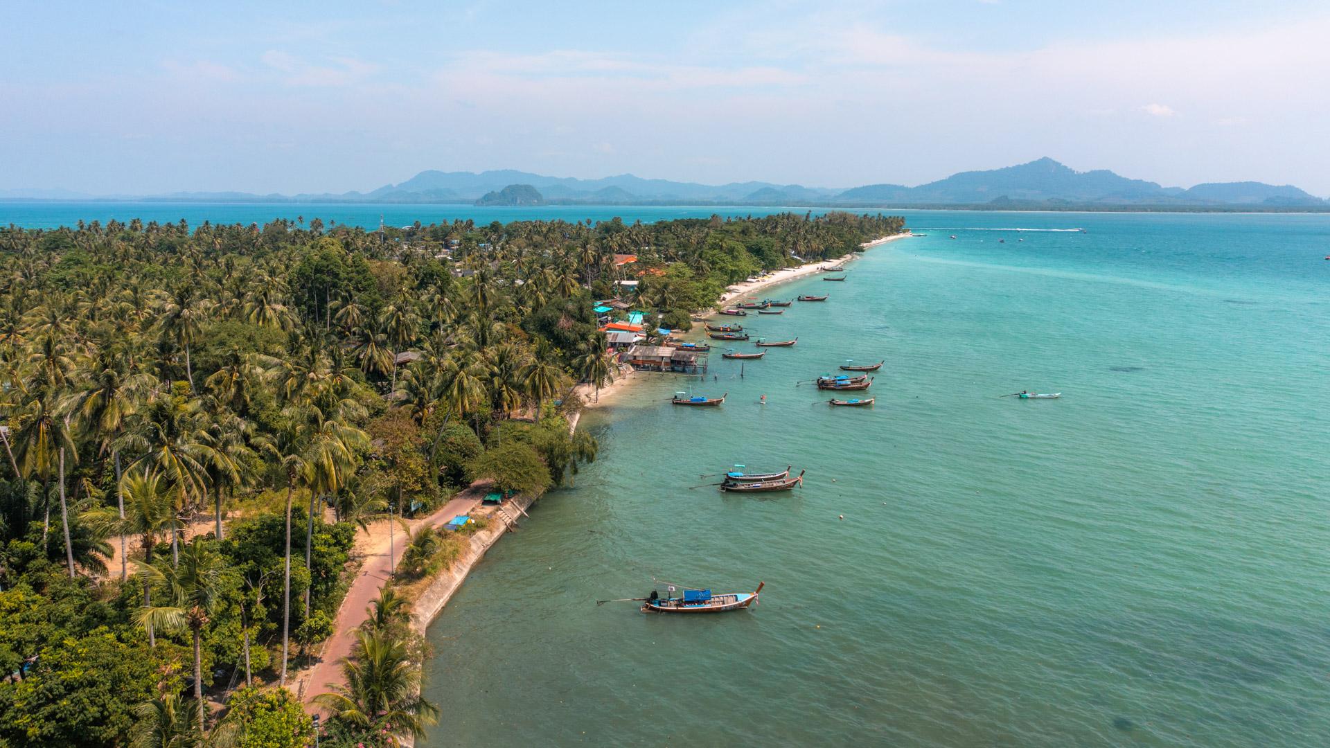 Aerial view of Koh Mook island showing turquoise waters, traditional longtail boats, palm trees and distant islands under clear sky.