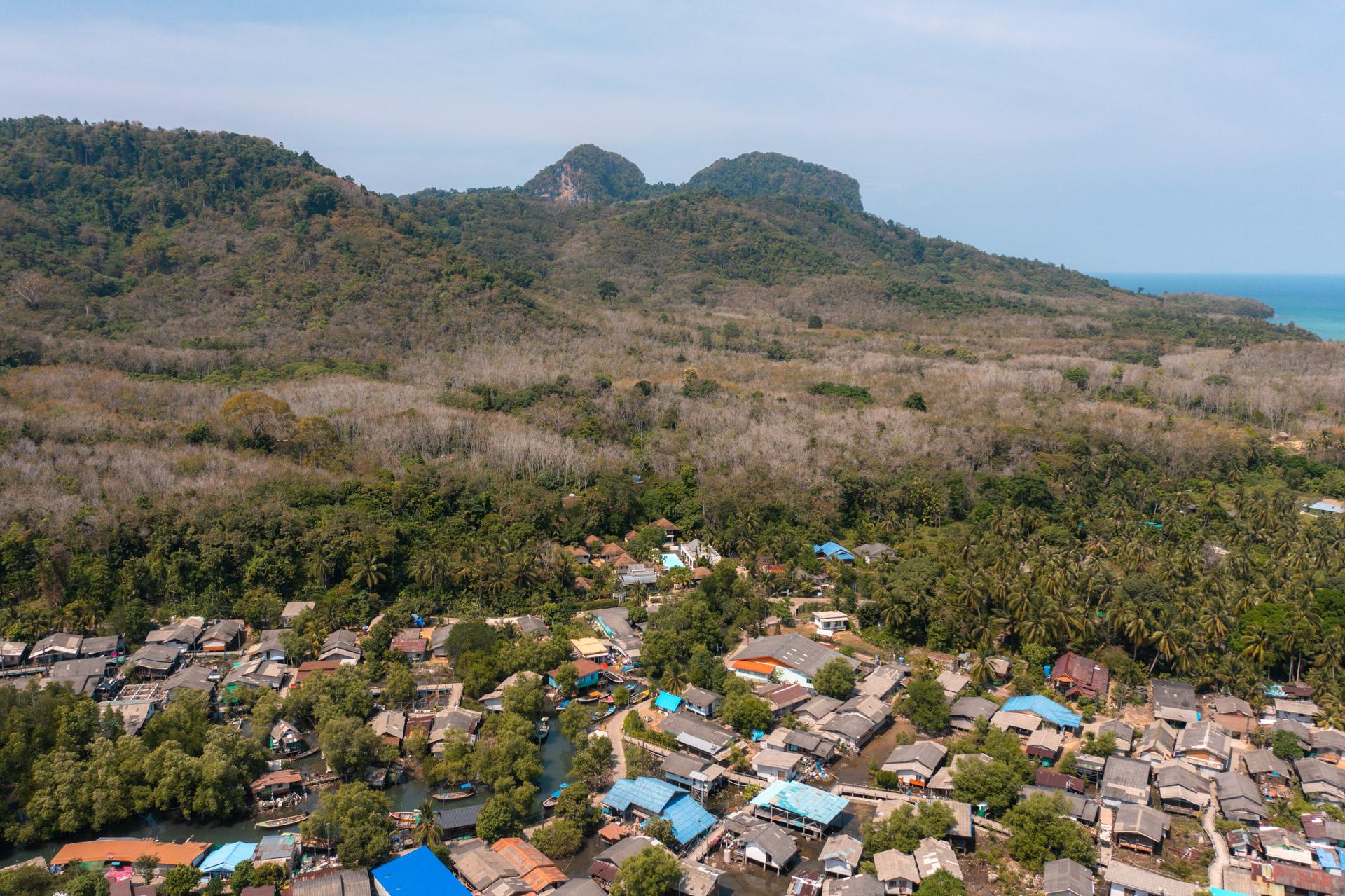 Aerial view of Koh Mook's village among palm trees, with forested hills and the turquoise sea visible beyond.