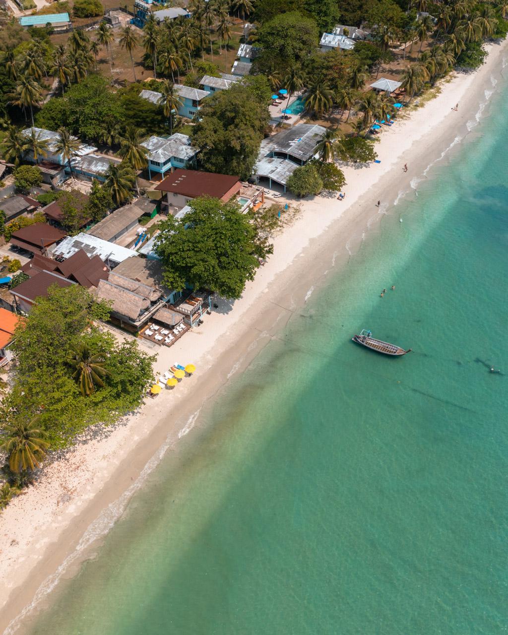 Aerial view of Koh Mook beach resort with palm trees, traditional buildings, turquoise water and wooden boat offshore.