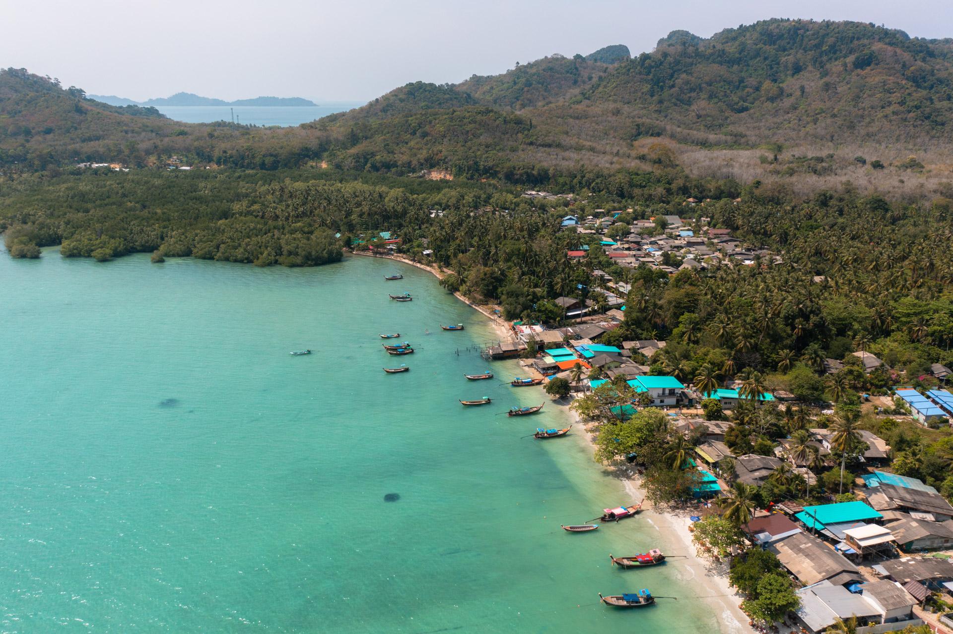 Aerial view of Koh Mook island with turquoise waters, longtail boats, coastal village with blue roofs, palm trees and forested hills.