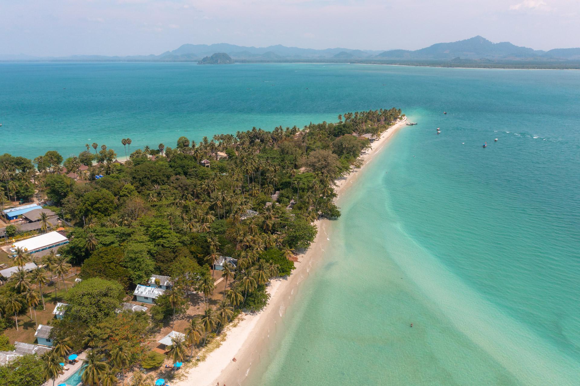 Aerial view of Koh Mook island with turquoise waters, white sand beach, dense palm trees and distant mountains.