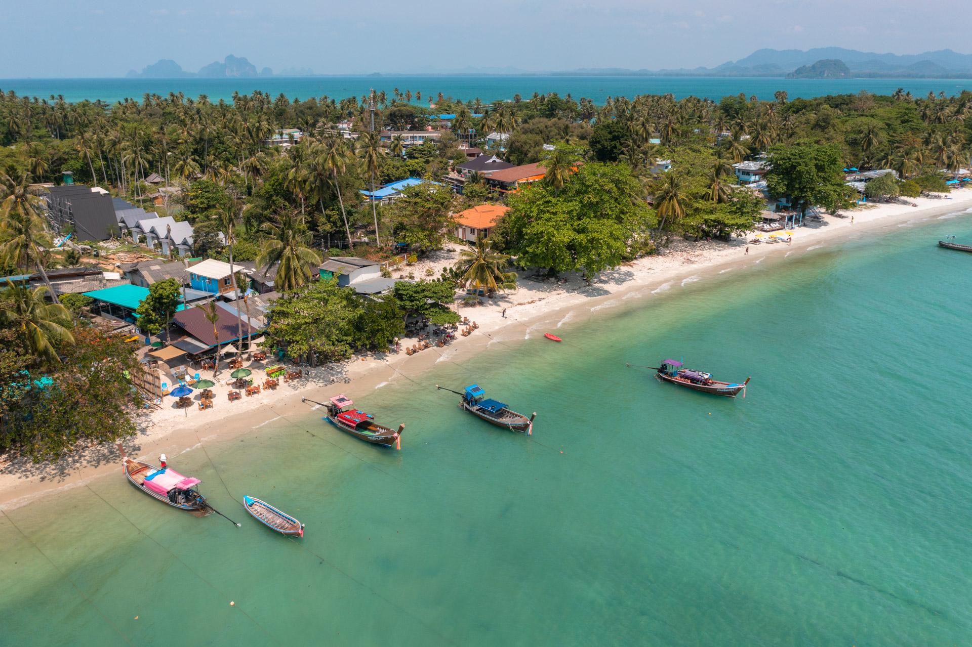 Low aerial view of Koh Mook beach with longtail boats moored offshore and a few low-rise places to stay tucked behind palm trees.