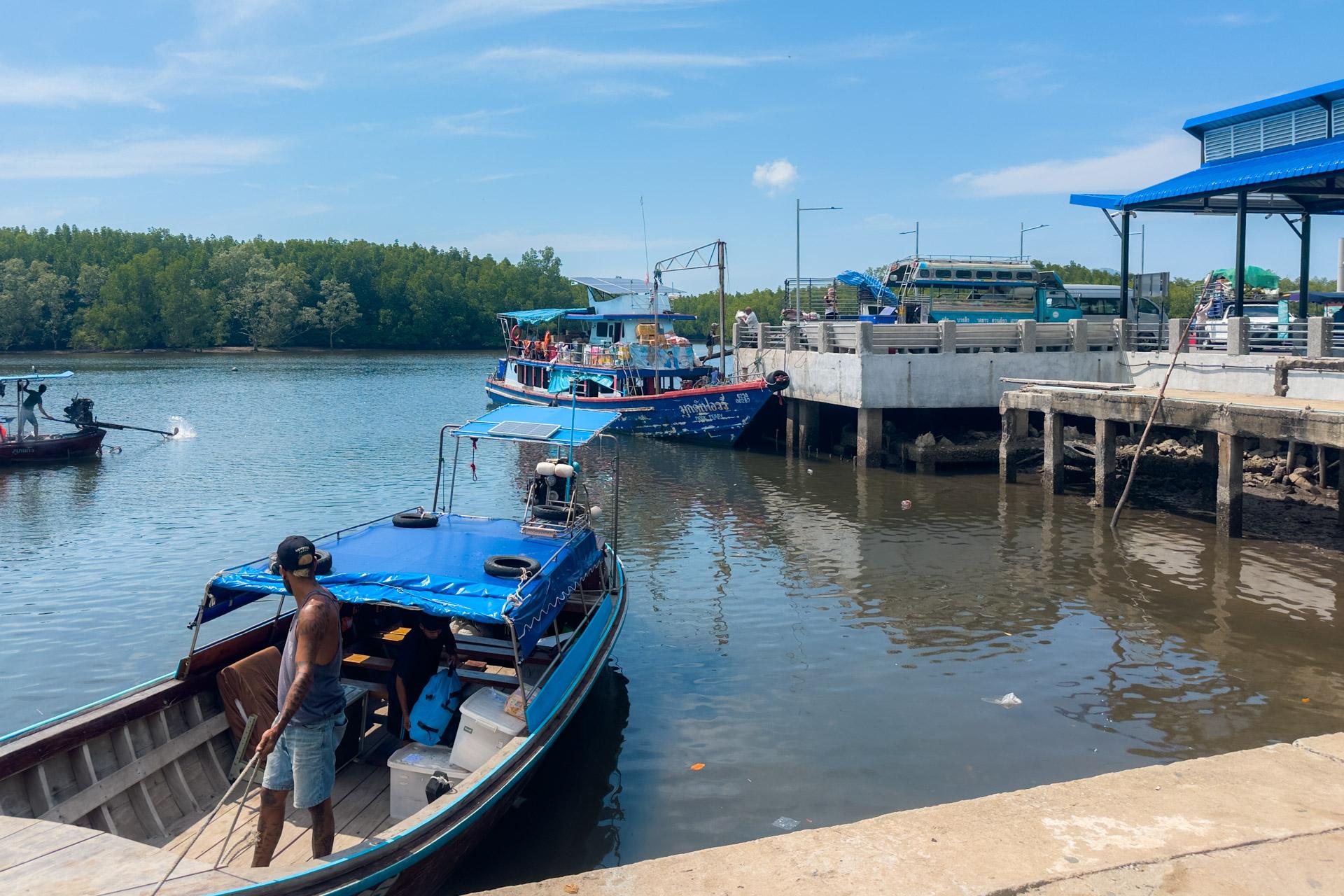 Blue and red fishing boats docked at Kuan Tung Ku Pier with mangrove forest and ferry terminal visible behind.