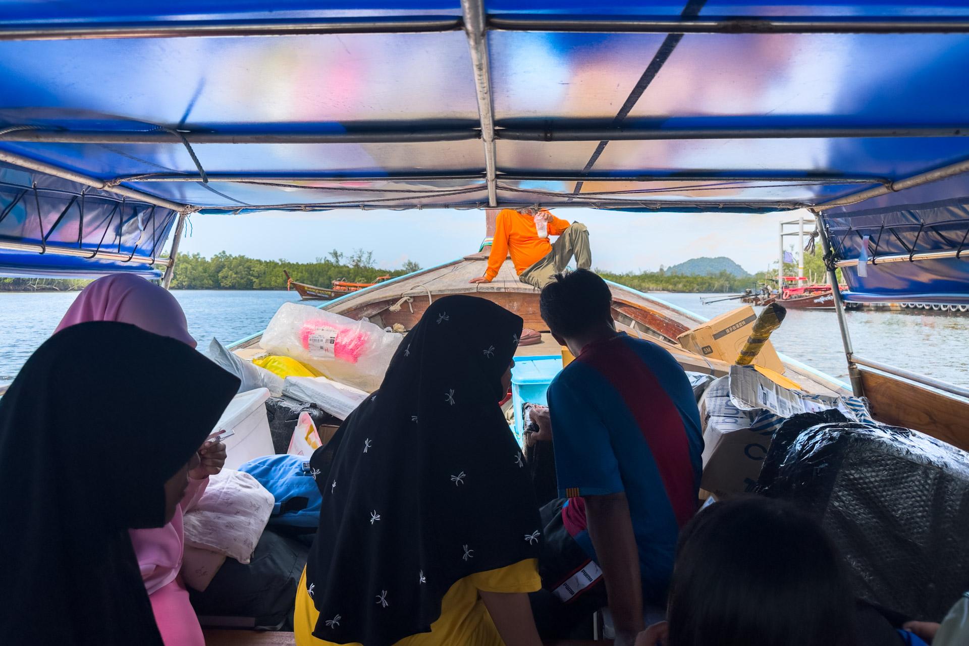 Passengers sit under blue canopy on traditional wooden longtail boat near Koh Mook, Thailand, with boatman and supplies visible.