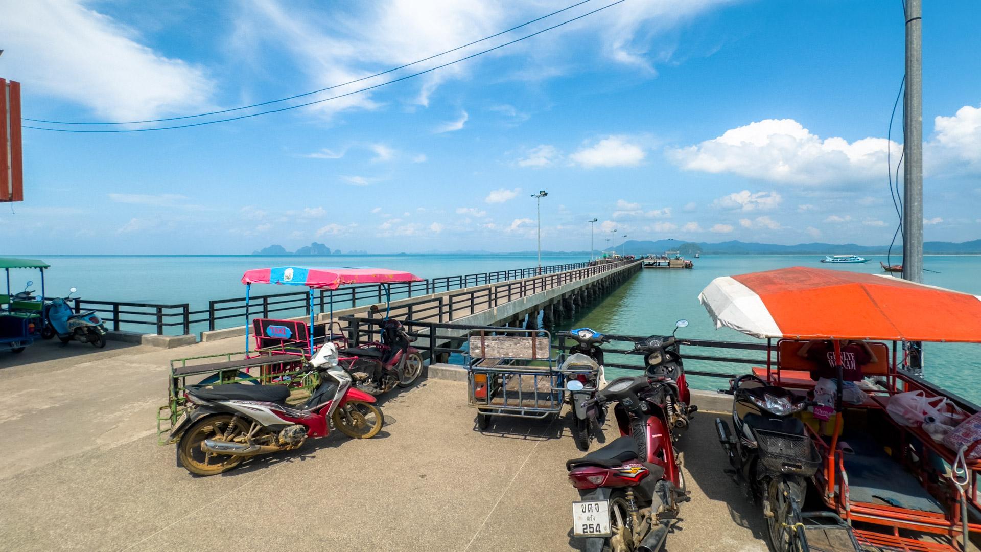 Koh Mook pier with colourful umbrellas, parked motorcycles, wooden pier extending into turquoise water and distant island mountains.