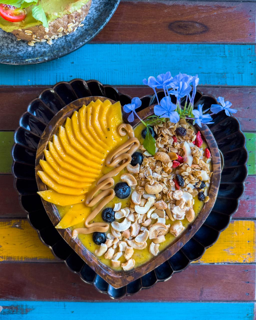 Heart-shaped smoothie bowl with sliced banana, granola, blueberries, cashews and blue flowers on colourful wooden surface in Koh Mook, Thailand.