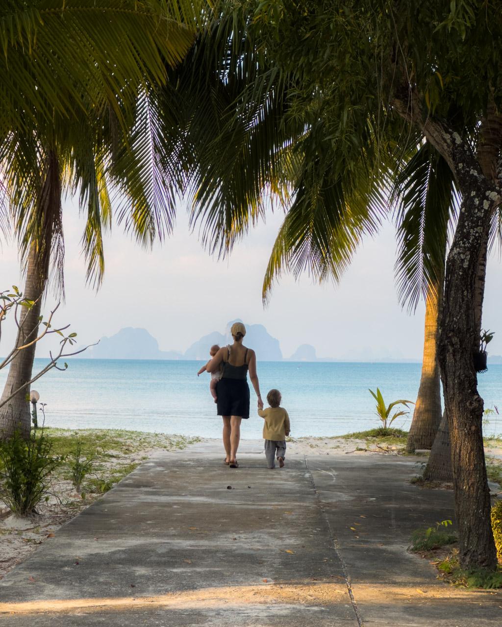 Victoria and two children walking on concrete path toward turquoise ocean between palm trees on Koh Mook beach, Thailand.