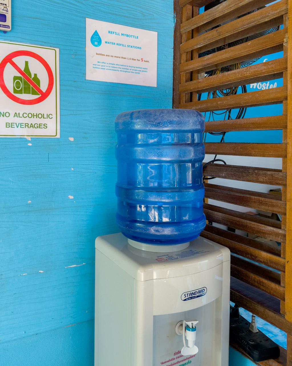 Water refill station with blue jug on dispenser, no alcohol sign on bright blue wall, Koh Mook, Thailand.