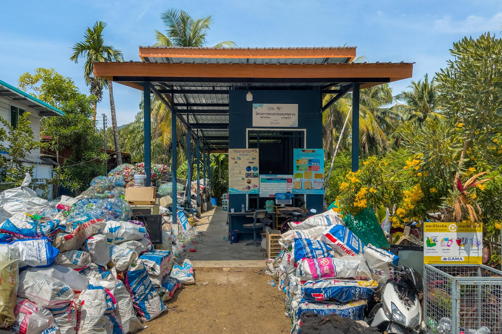Community recycling centre on Koh Mook with sorted plastic waste bags and educational signage under a wooden structure.