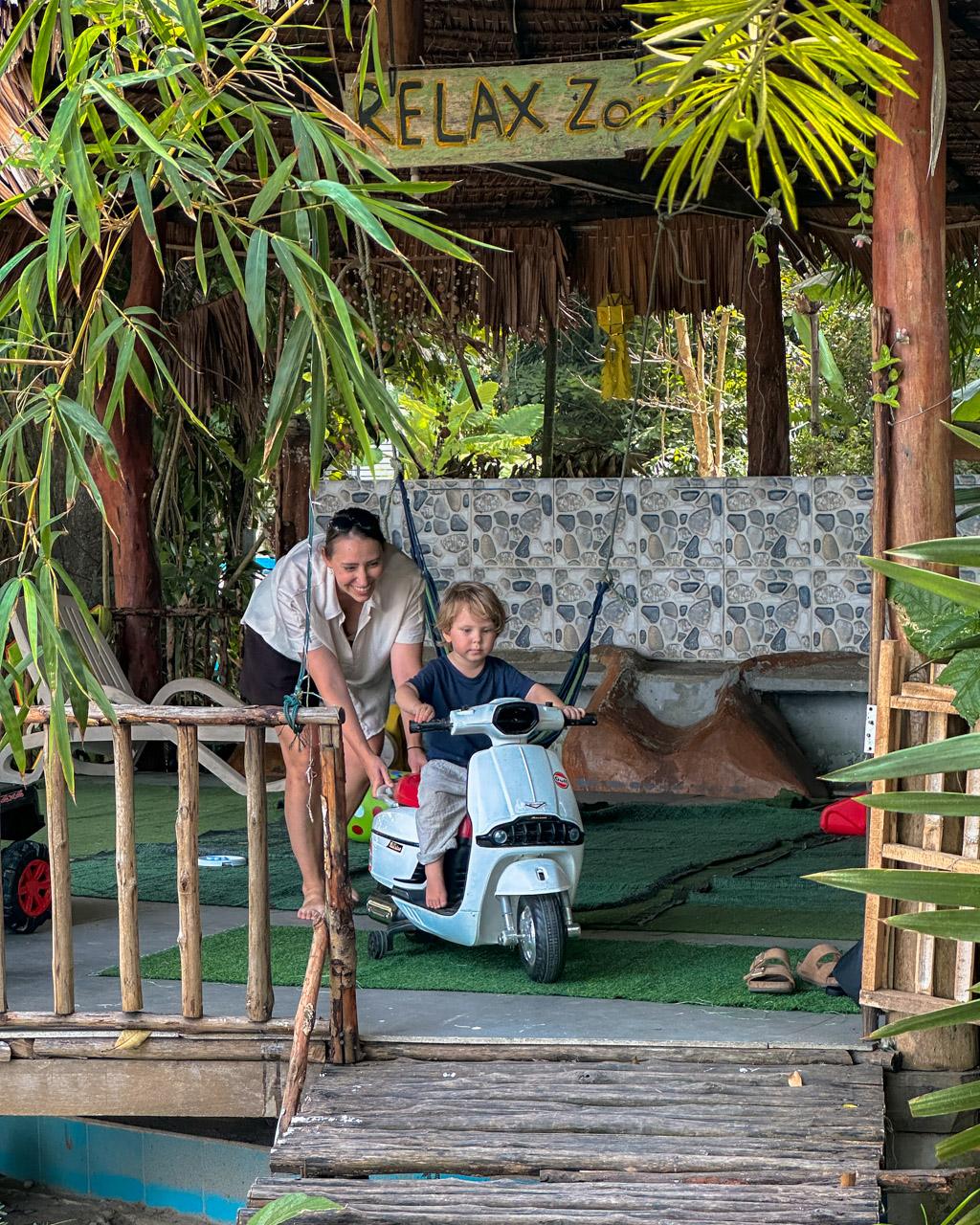 Adult and child on white toy scooter under "Relax Zone" shelter with tropical plants, Koh Mook Thailand.