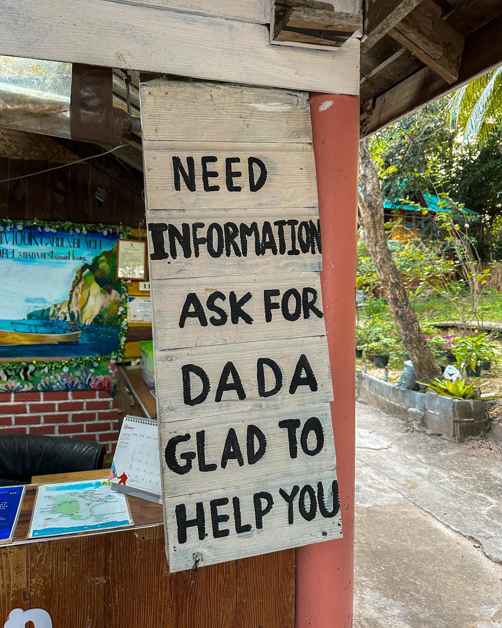 Wooden sign at Koh Mook reading "Need information ask for Dada glad to help you" with tropical foliage visible.