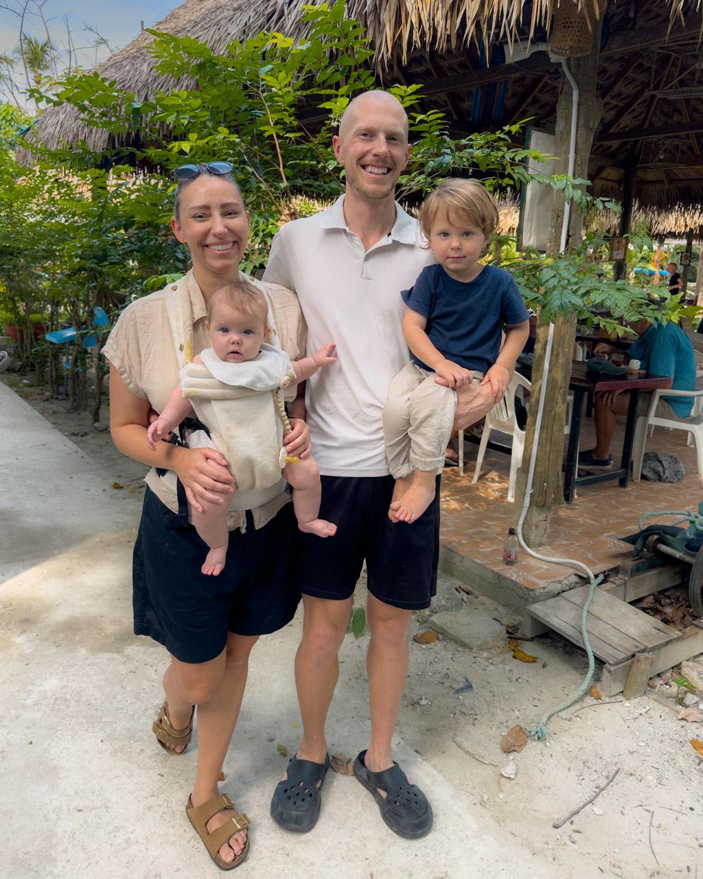 Family of four smiling at tropical beach bungalow in Koh Mook, Thailand, with thatched roof structures behind them.