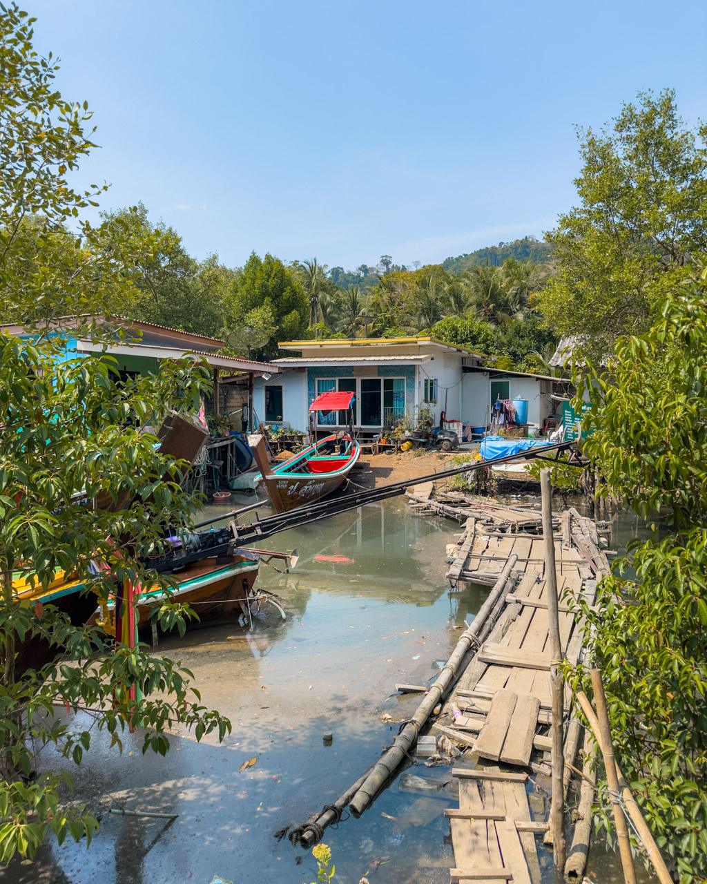 Wooden walkway over mangrove canal leading to blue and white fishing village house with colourful boats moored in shallow water, Koh Mook, Thailand.