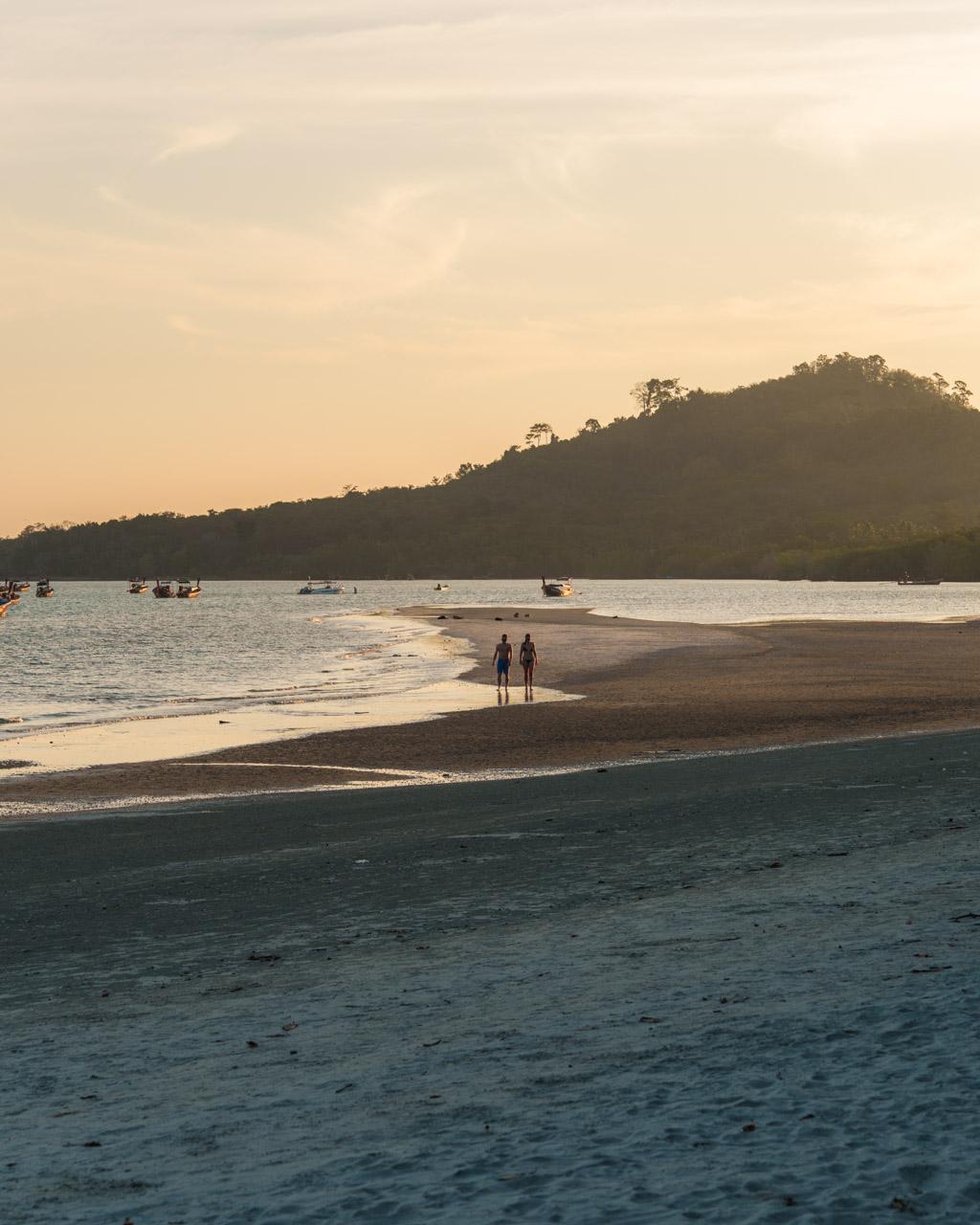 Two people standing in shallow water at Koh Mook beach at sunset, with forested hillside and moored boats in background.