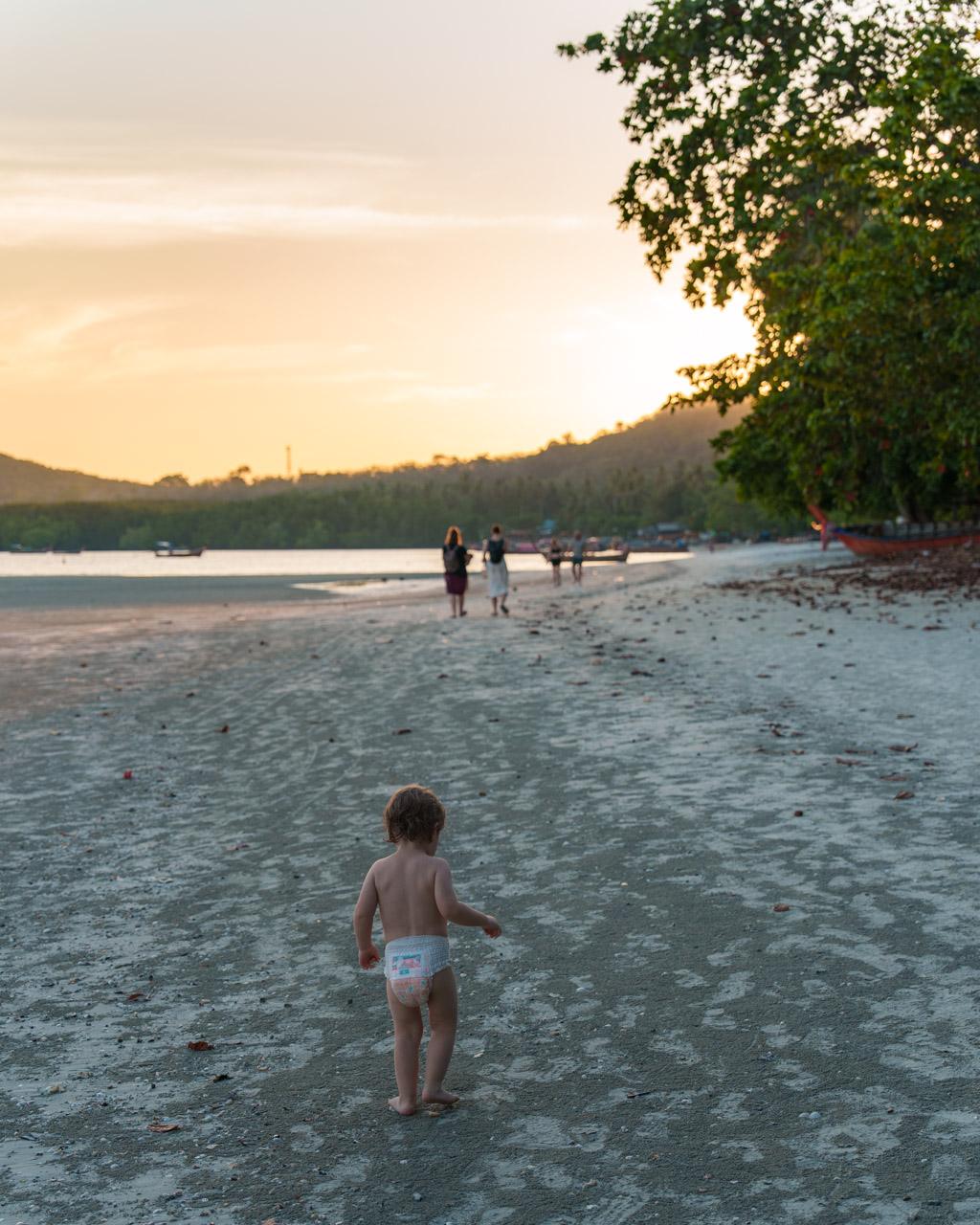 Young child wading on sandy beach at sunset in Koh Mook, Thailand, with lush vegetation and boats in background.