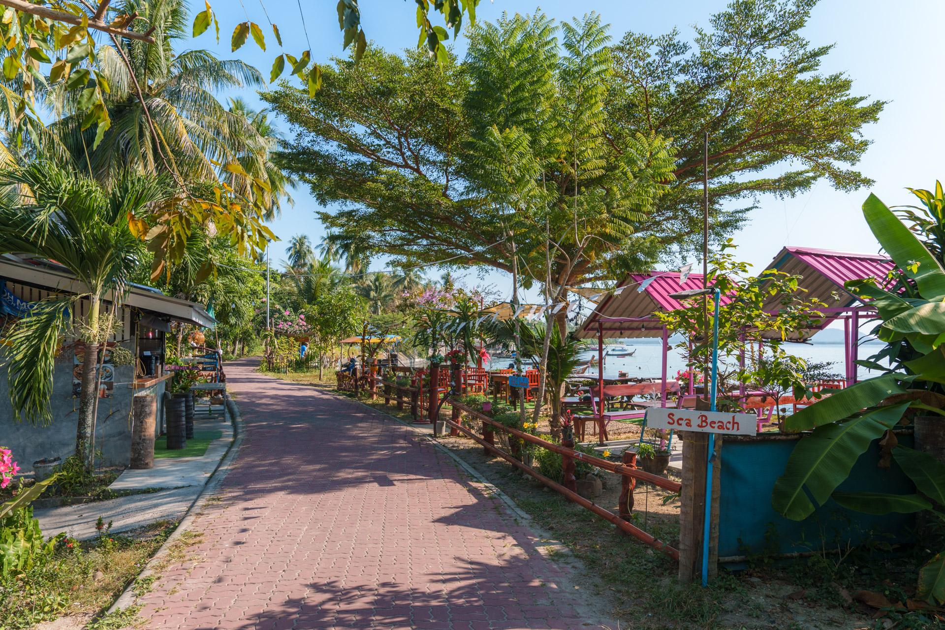 Tropical beach pathway in Koh Mook, Thailand with pink-roofed pavilions, palm trees and a sign for Sea Beach Restaurant.
