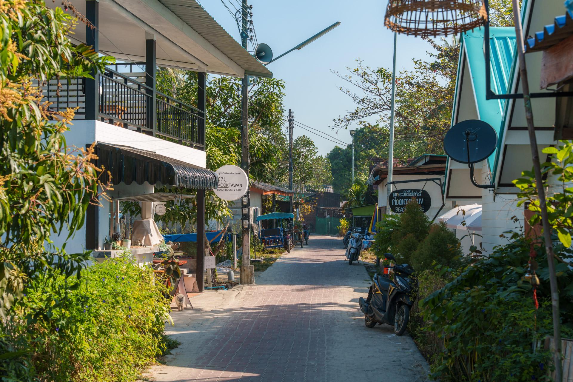 Quiet village street in Koh Mook, Thailand, with shops, motorbikes, lush vegetation and tropical buildings under a clear sky.