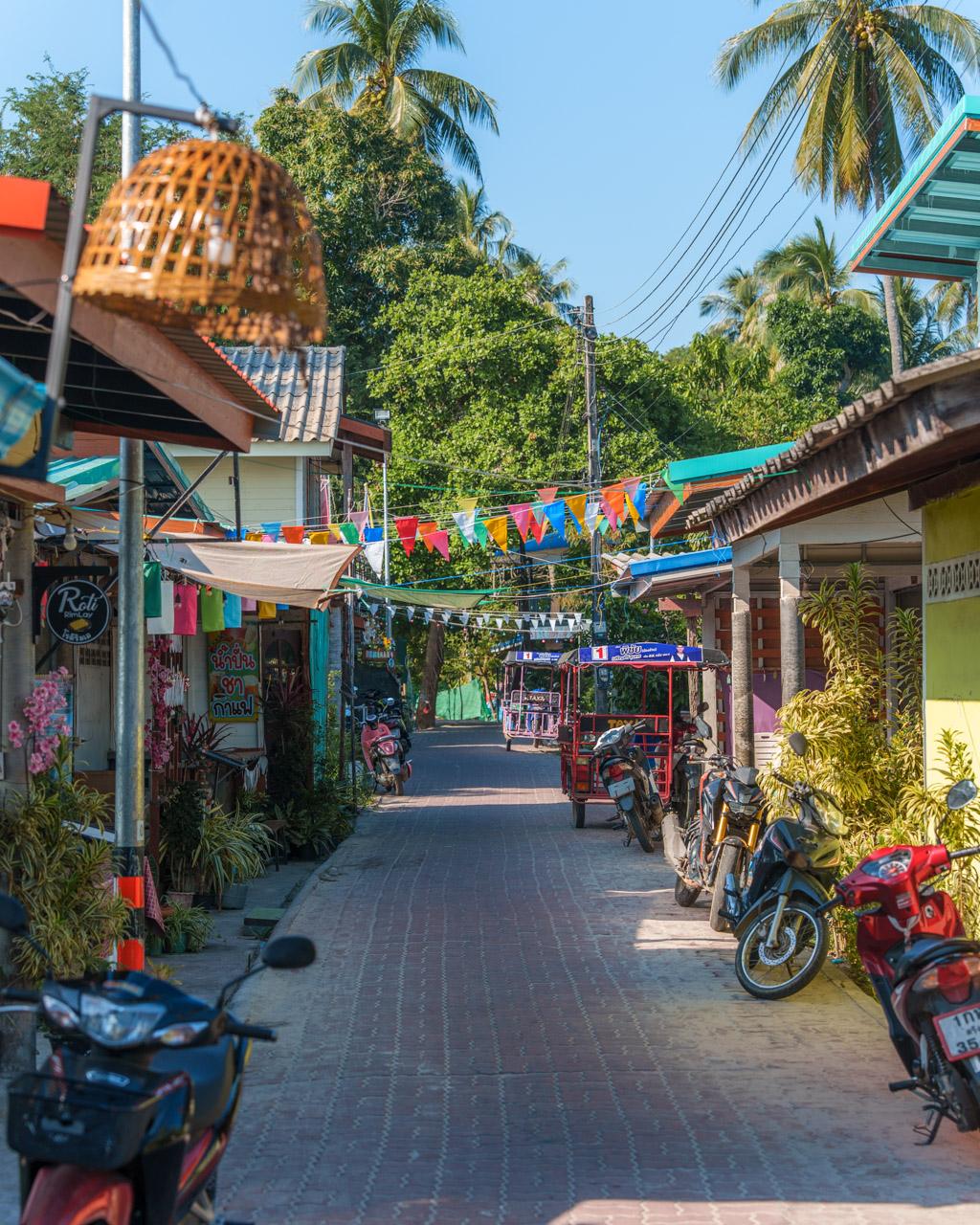 Colourful village street in Koh Mook, Thailand with shops, motorcycles, palm trees and festive bunting decorations.