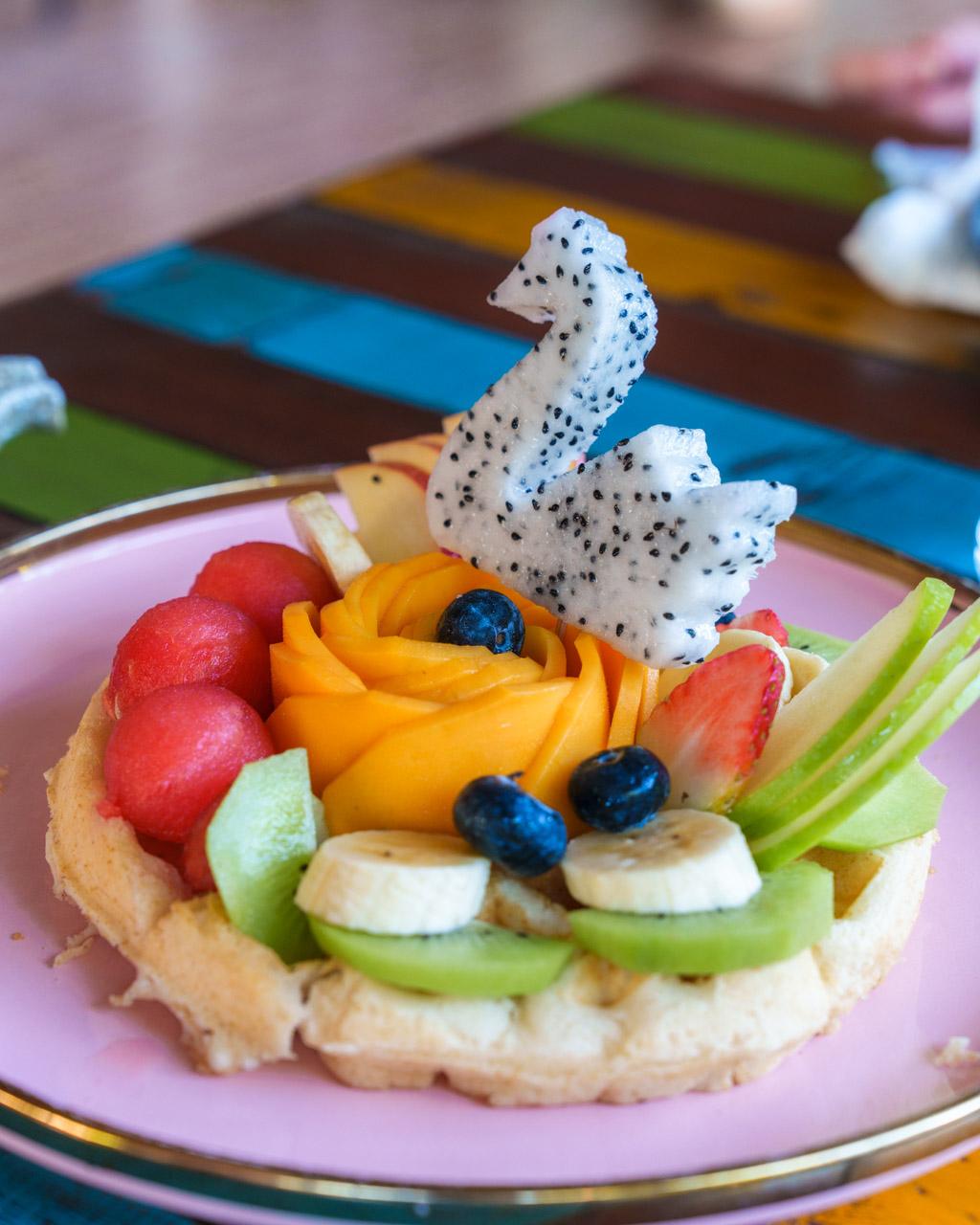 Artfully plated vegan waffle topped with dragon fruit, mango, strawberries, blueberries, kiwi and banana on pink plate.