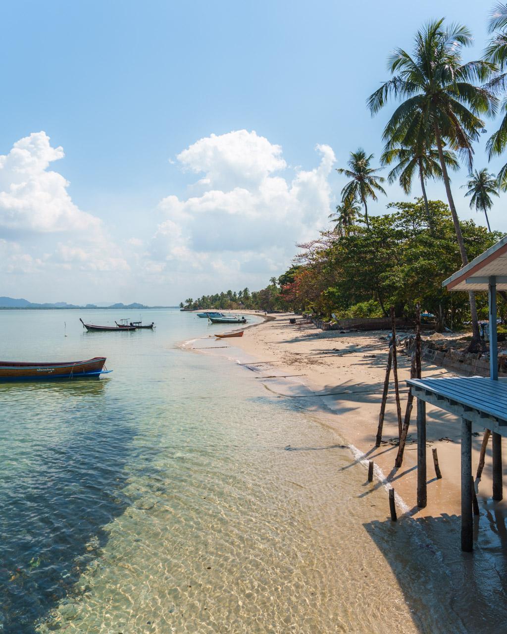 Koh Mook tropical beach with clear turquoise water, wooden pier, moored longtail boats, palm trees and blue sky with white clouds.