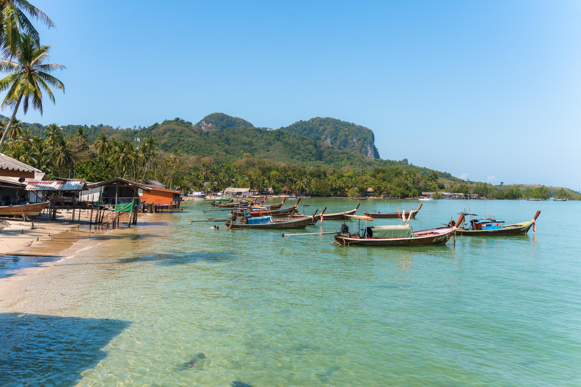 Koh Mook beach with traditional longtail boats, palm trees, and forested limestone hills under a clear blue sky.