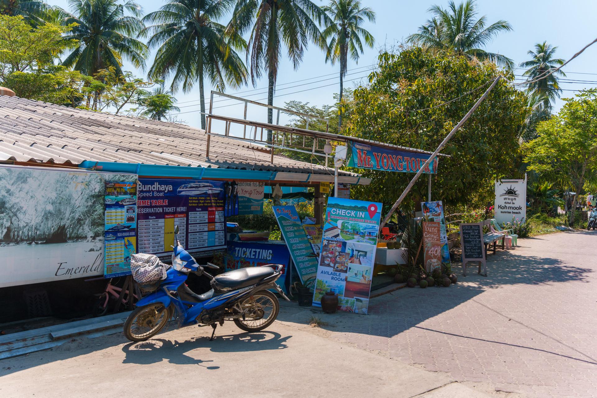 Koh Mook beachfront ticket booth with blue signage, speedboat tours, parked scooter and palm trees.