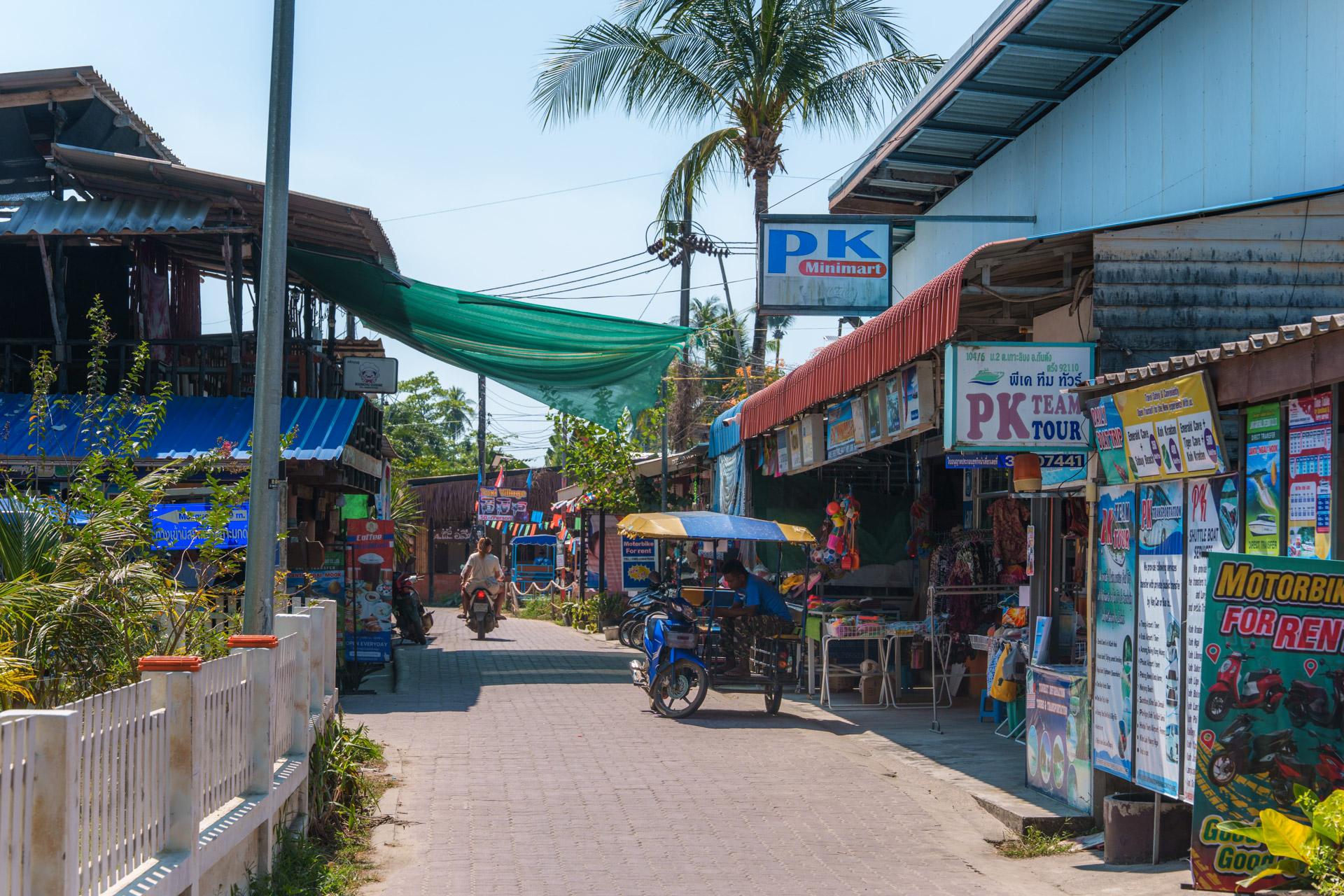Busy street market in Koh Mook, Thailand, with shops, motorbikes, a palm tree and colourful signage under a blue sky.