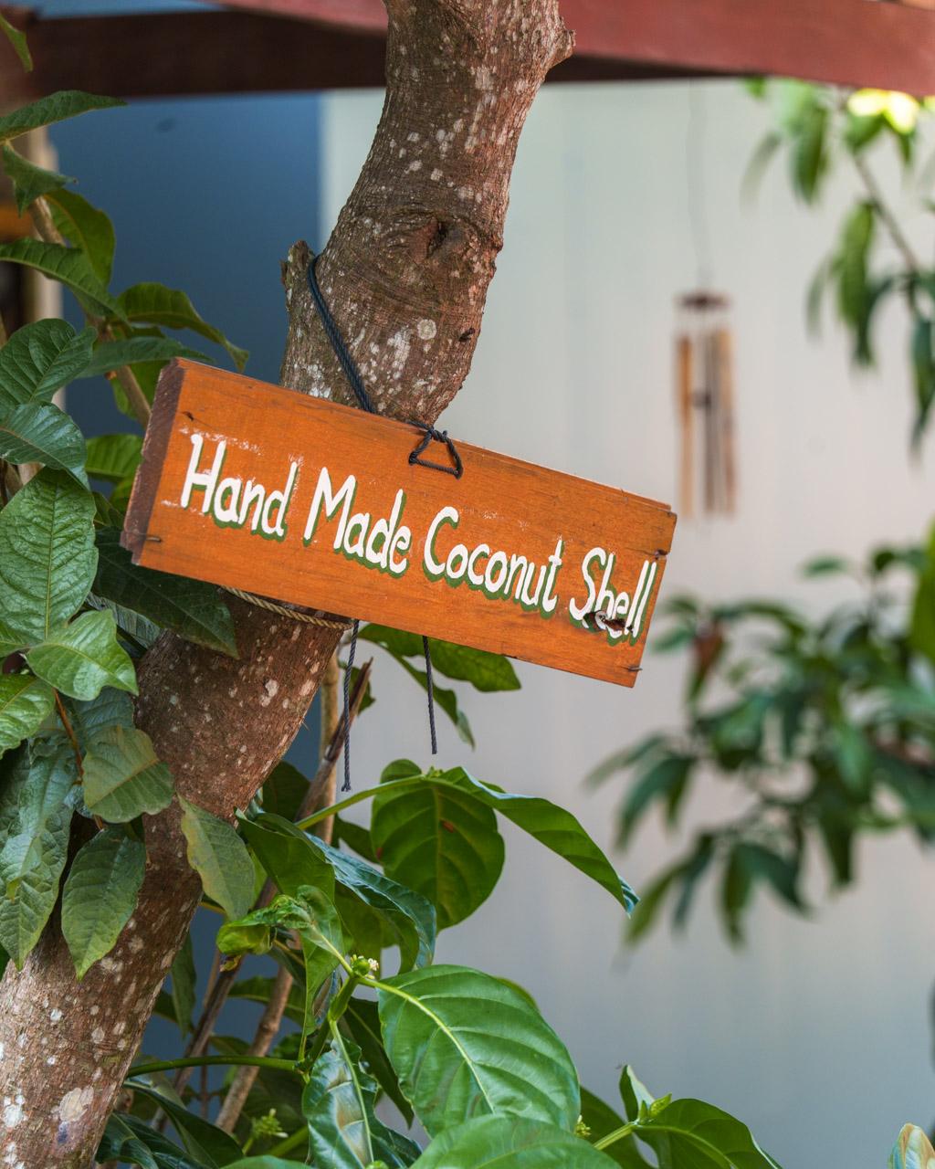 Orange wooden sign reading "Hand Made Coconut Shell" hanging from tree branch with green leaves in Koh Mook, Thailand.