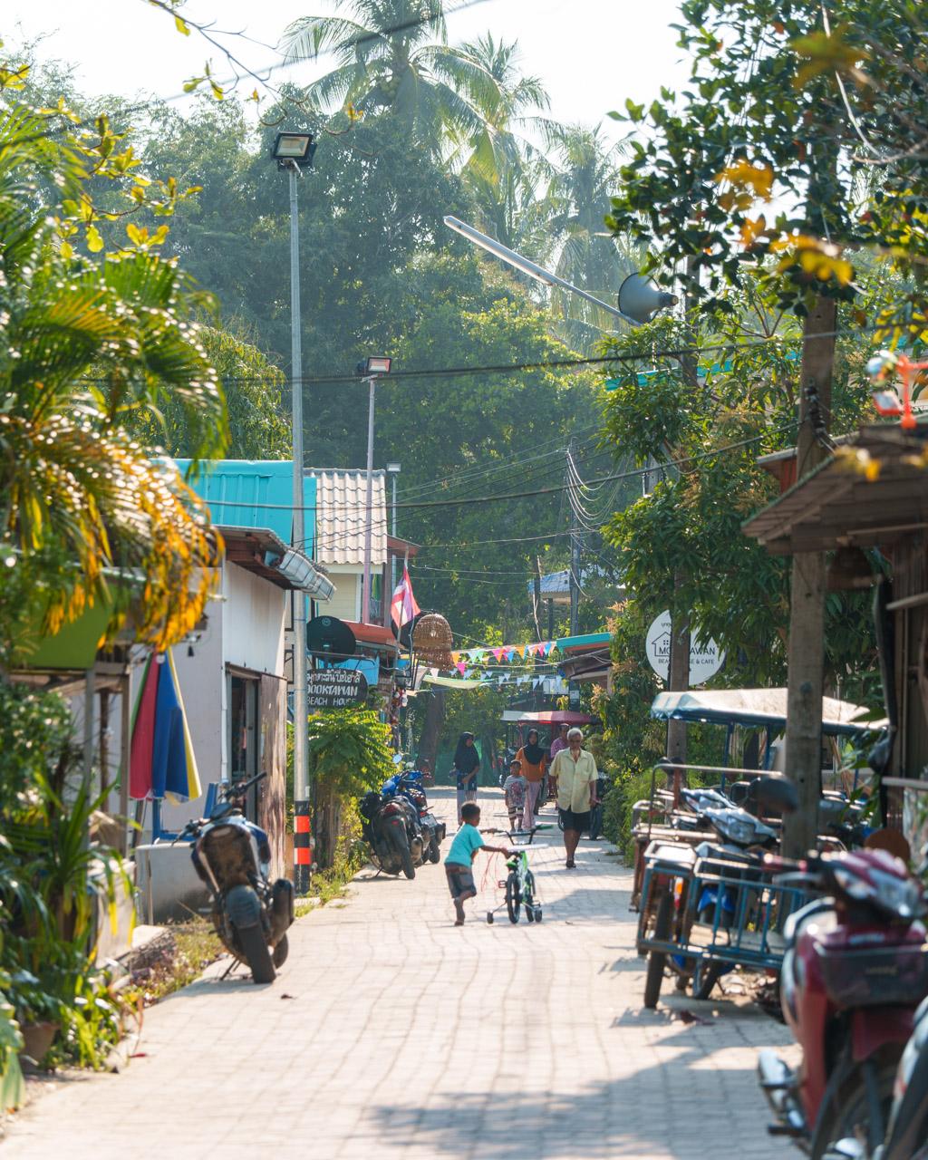 Tropical village street in Koh Mook, Thailand, with colourful flowers, palm trees, local shops, motorcycles and residents walking.