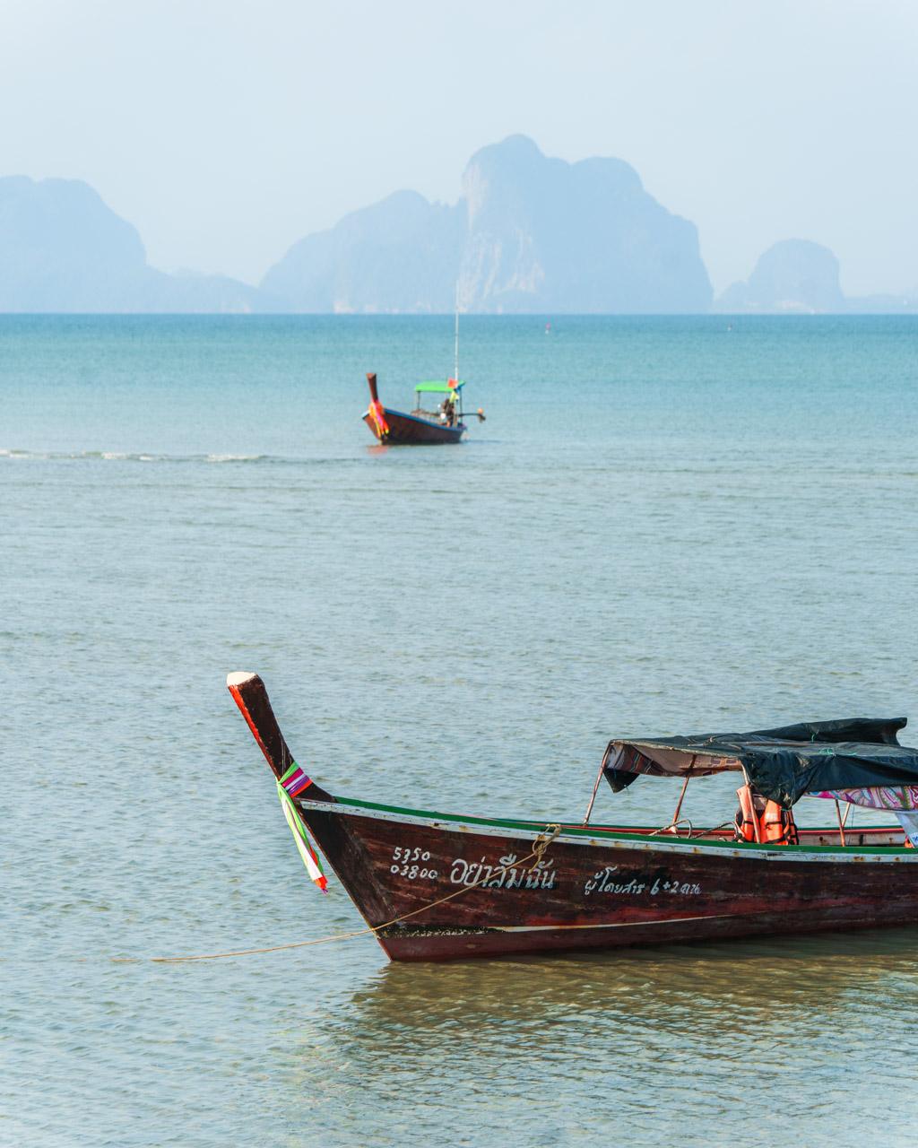 Traditional wooden longtail boats anchored in turquoise water off Koh Mook, Thailand, with limestone cliffs in the background.