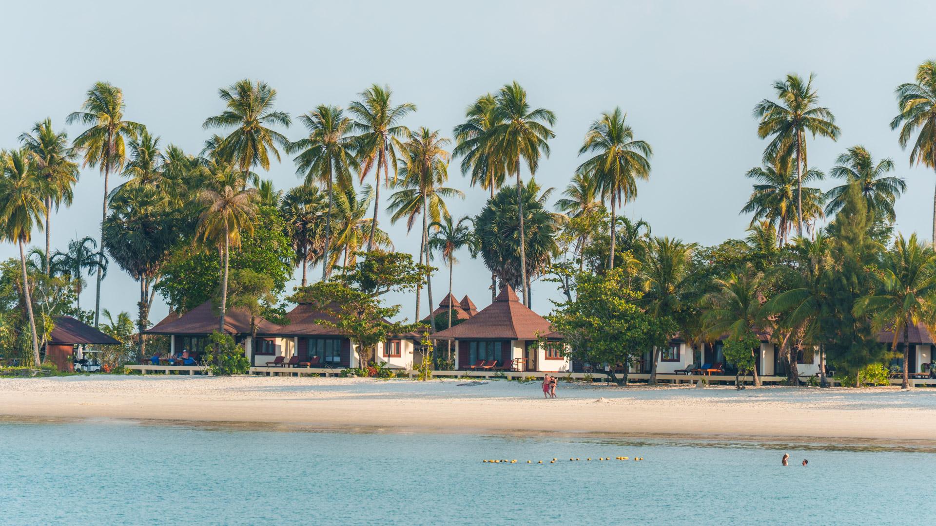 Tropical beachfront resort (Sivalai) with red-roofed bungalows, tall palm trees, sandy beach and shallow turquoise water on Koh Mook island, Thailand.