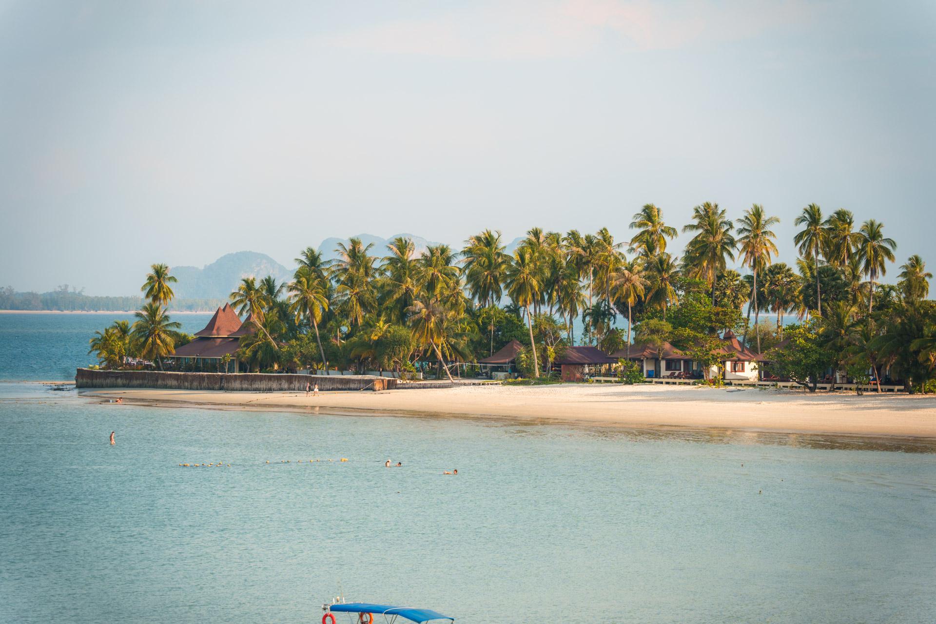 Tropical beach resort on Koh Mook with palm trees, traditional wooden bungalows, sandy shore and swimmers in turquoise waters.