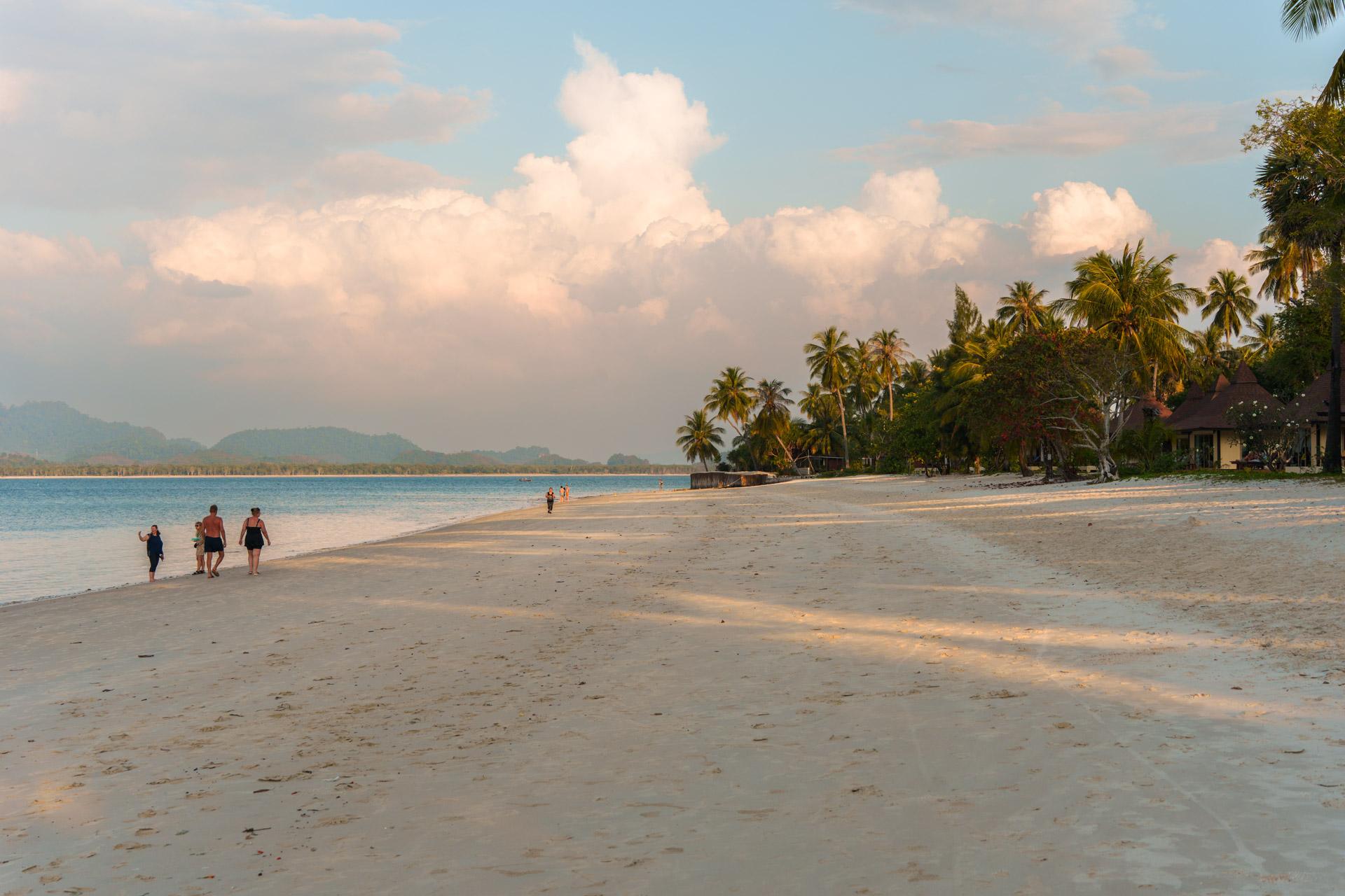 Tropical beach in Koh Mook, Thailand with turquoise water, palm trees, beachfront bungalows and swimmers under cloudy sky.