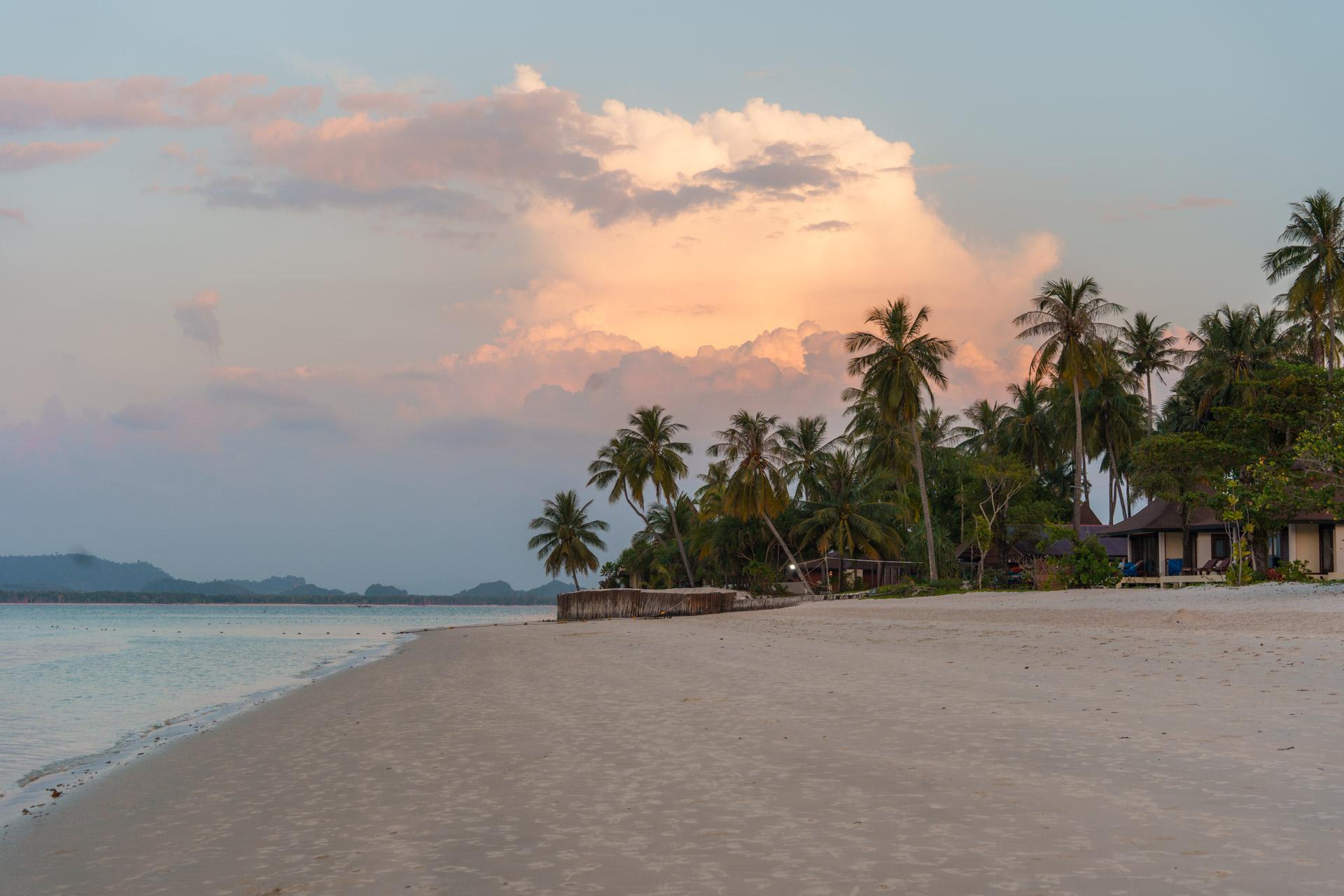 Koh Mook beach at sunset with golden clouds, palm trees, sandy shore, turquoise water and wooden bungalows along the coastline.