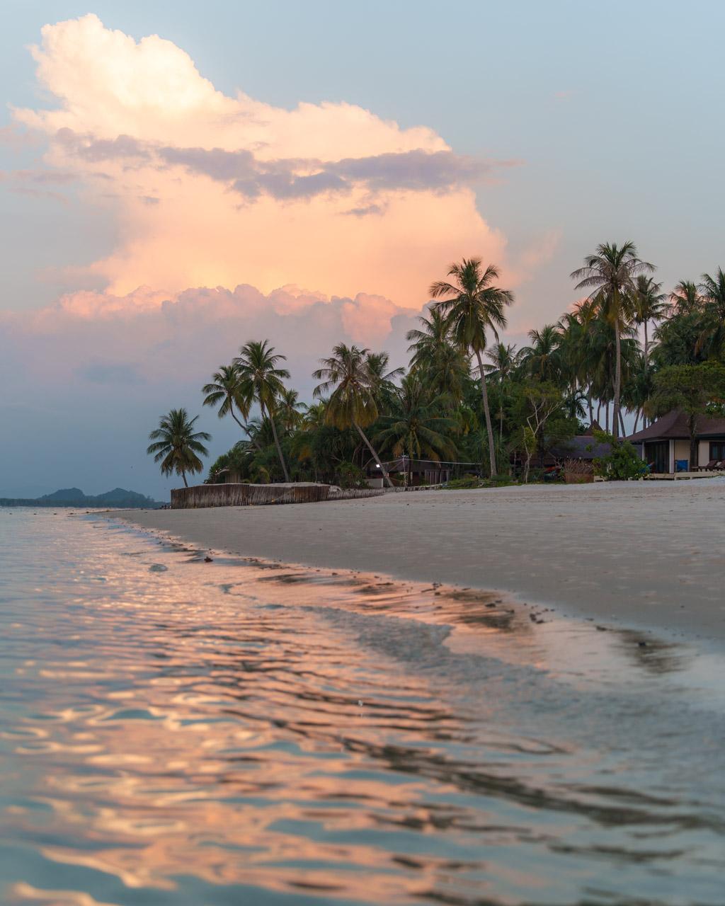 Tropical beach at sunset with palm trees, wooden structures and pink-tinted clouds over calm waters on Koh Mook island.