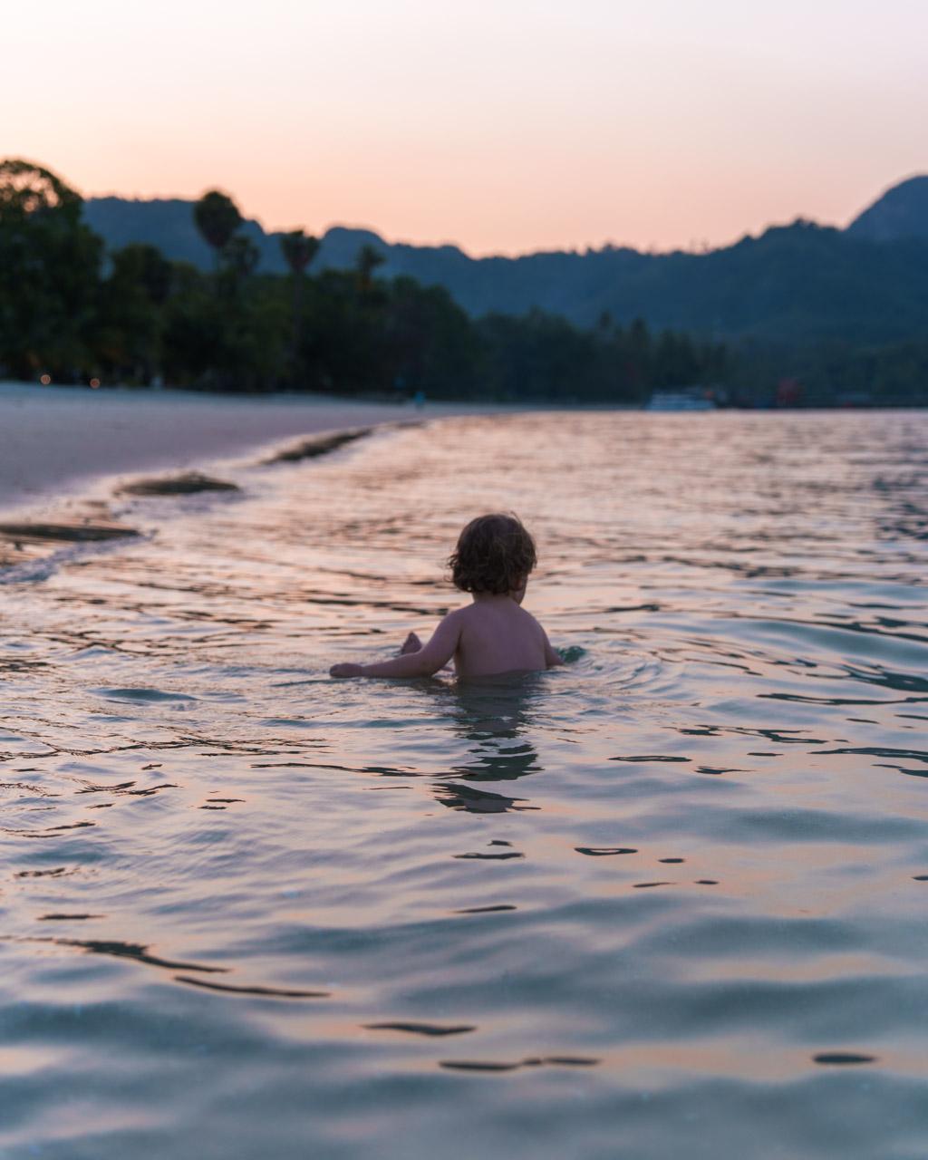 Child wading in shallow water at sunset on Koh Mook beach, forested limestone mountains in the background.