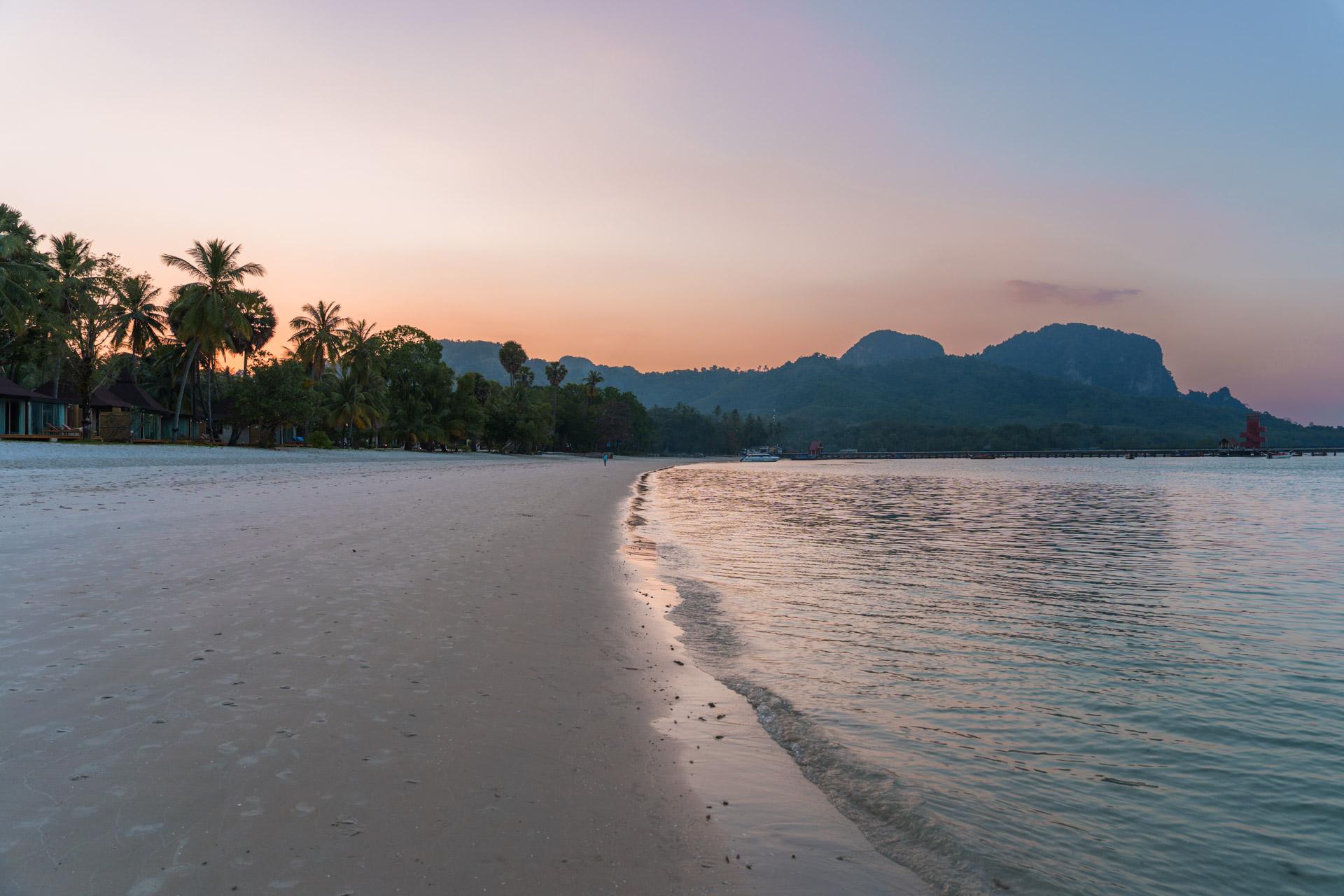 Sunset over Koh Mook beach with limestone cliffs, palm trees and calm turquoise waters at dusk.