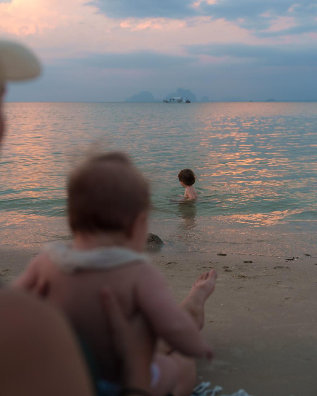 Child (Adrian) wading in shallow turquoise water at Koh Mook beach, with distant limestone islands and sunset sky reflected on water. In the foreground there's a child and mother blurred (Zoe and Victoria).