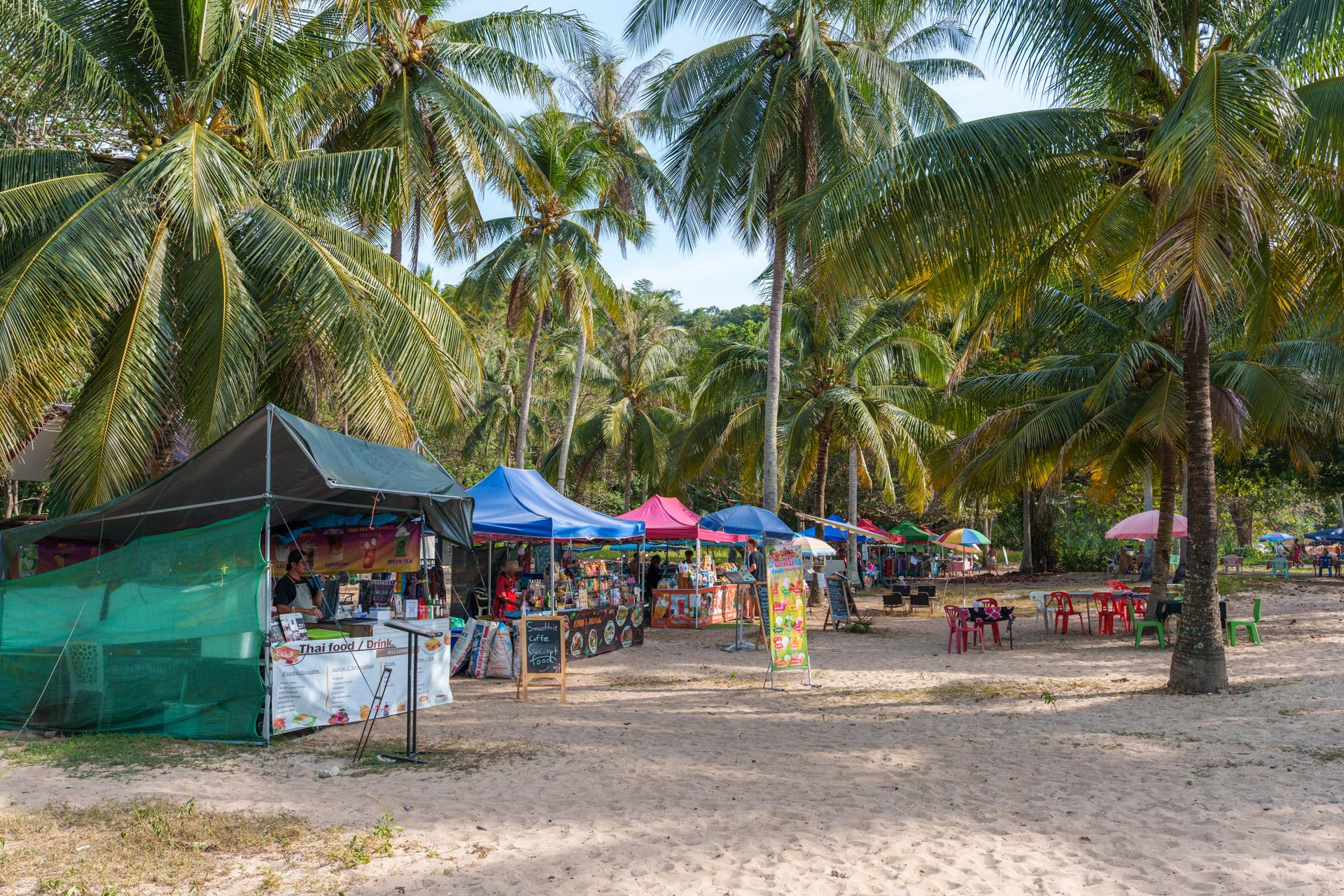 Beach market with colourful vendor tents and umbrellas surrounded by tall coconut palms in Koh Mook, Thailand.